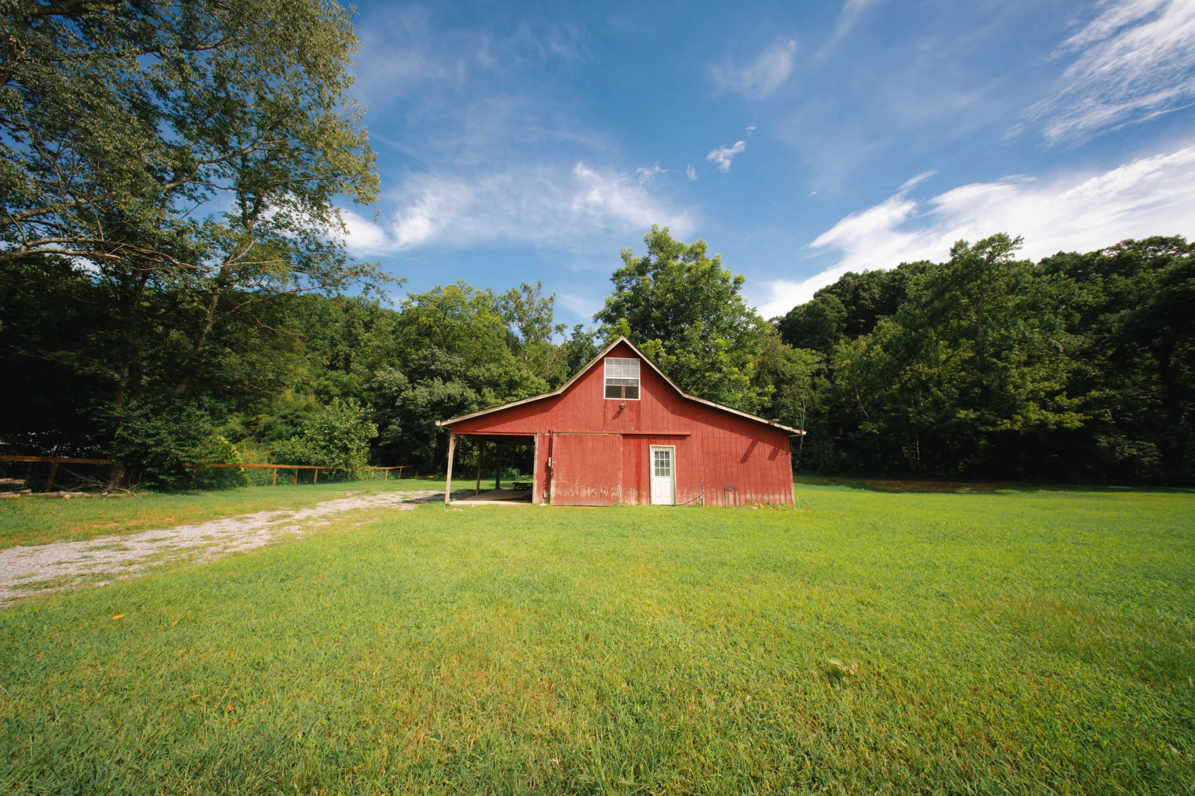 A red barn stands on a grassy area surrounded by trees under a blue sky.