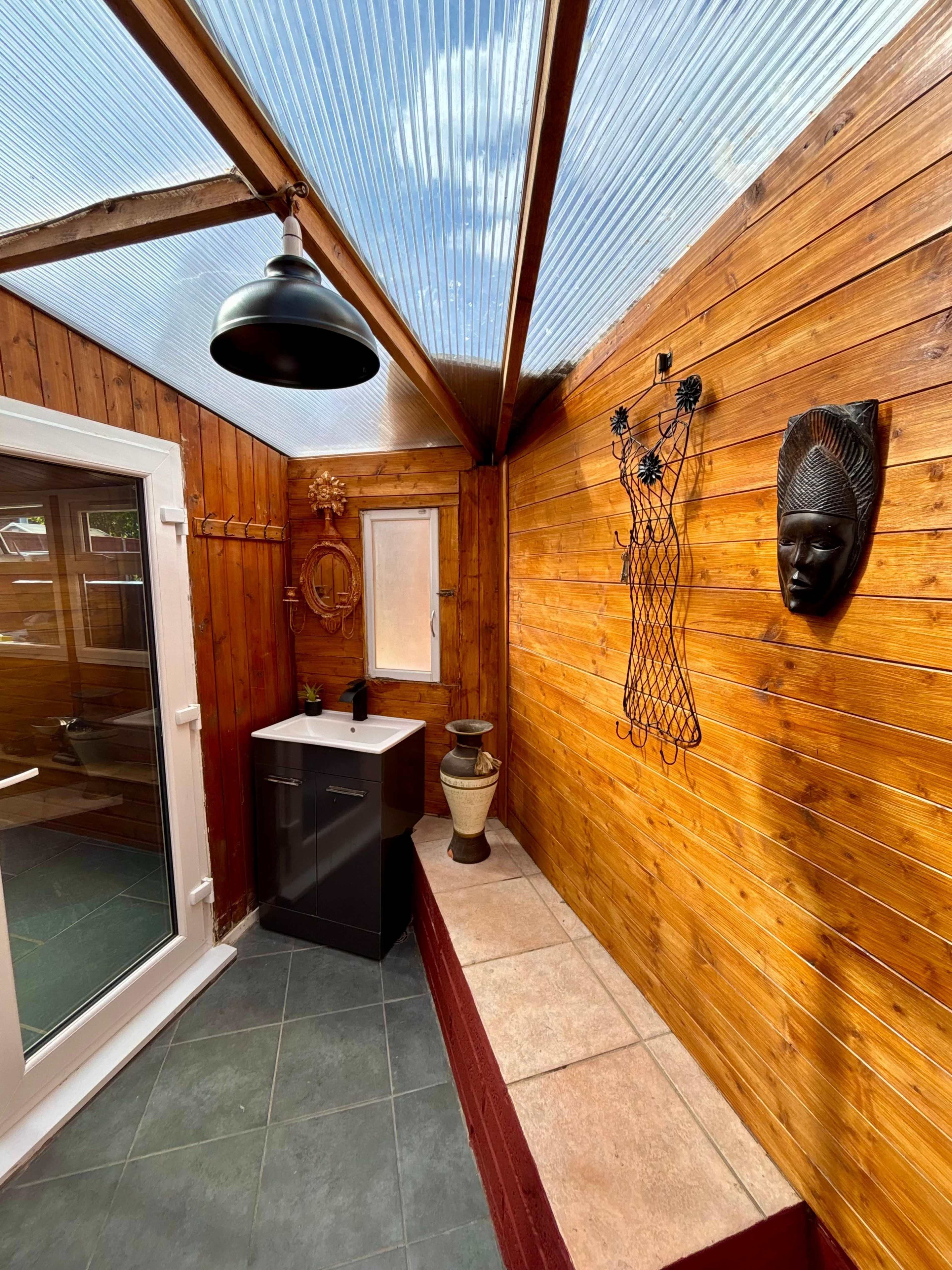 The image shows a wooden sunroom with a tiled floor, featuring a black sink unit, a decorative wall mask, and a large window allowing natural light inside.