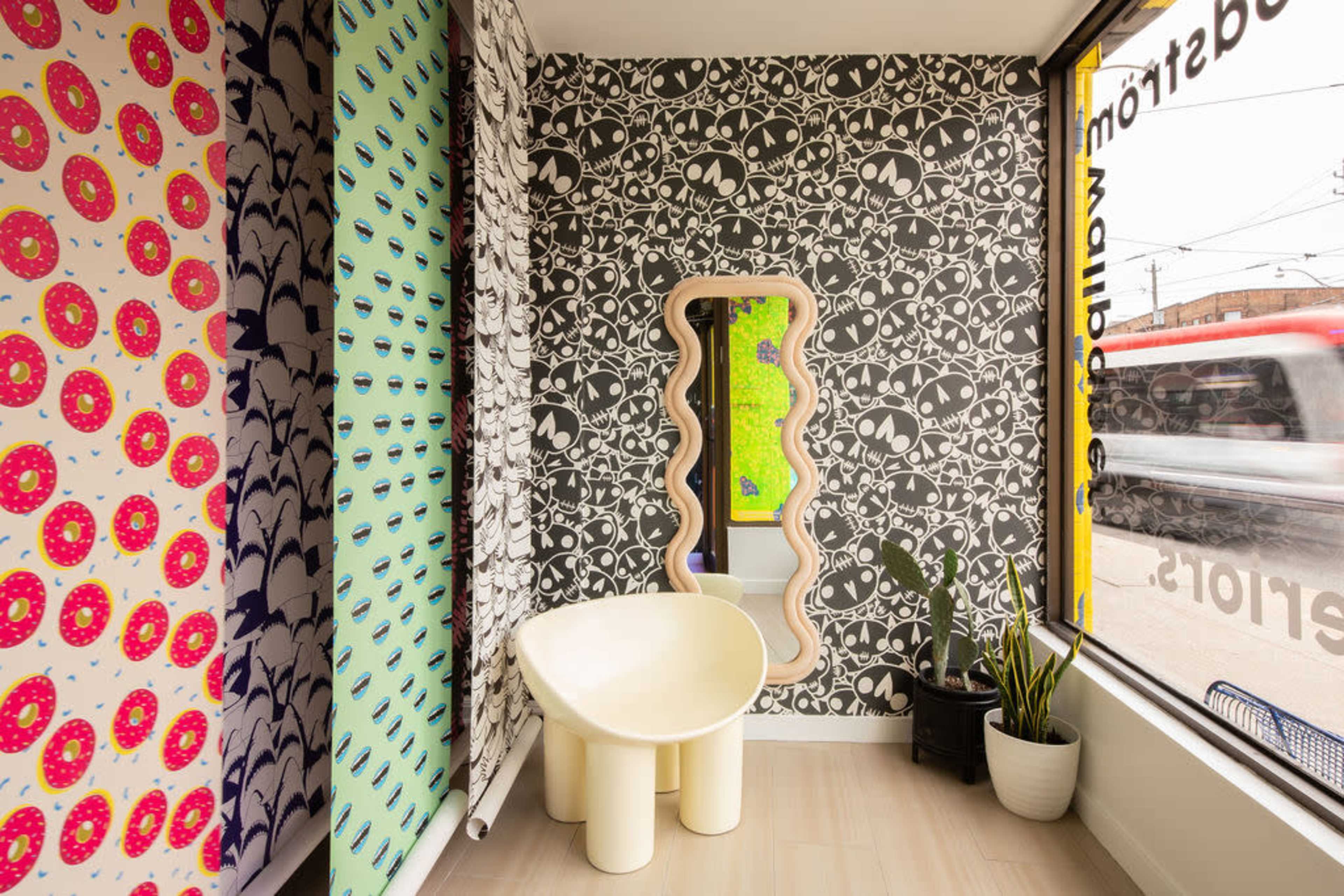 The image shows a brightly decorated corner of a room featuring patterned wallpaper and a curved mirror, with a unique yellow chair and a small plant by the window.