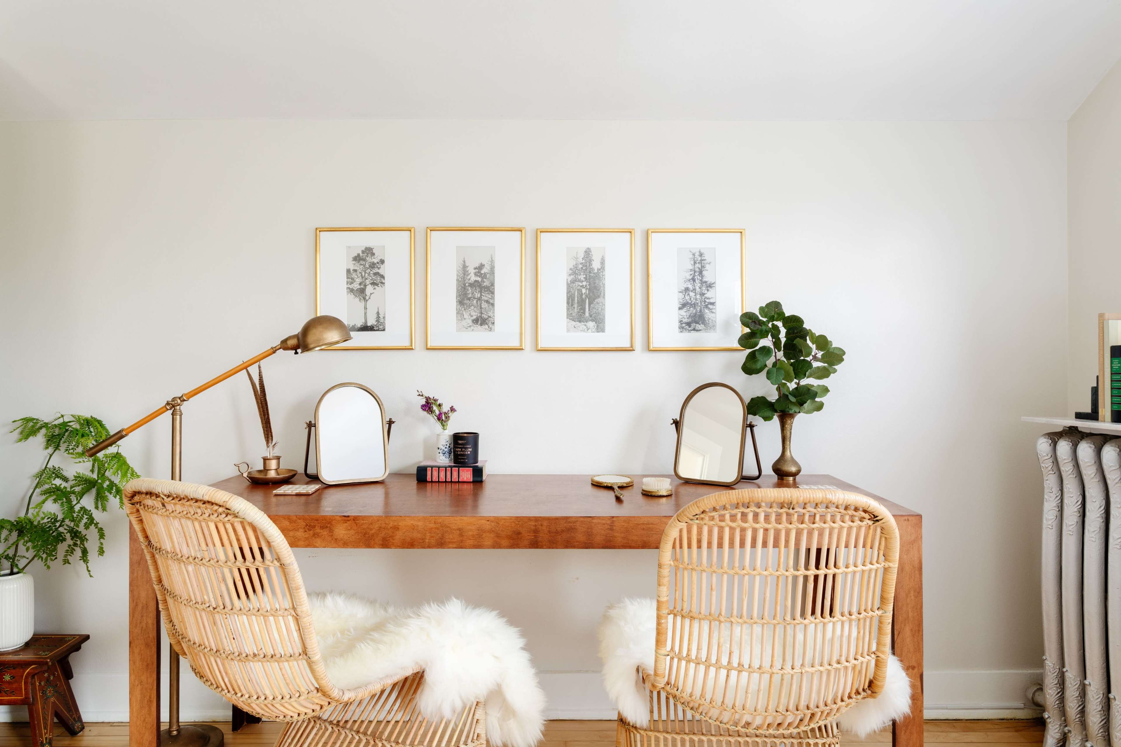 A wooden desk with two rattan chairs, adorned with framed nature prints and a potted plant, is positioned against a white wall.