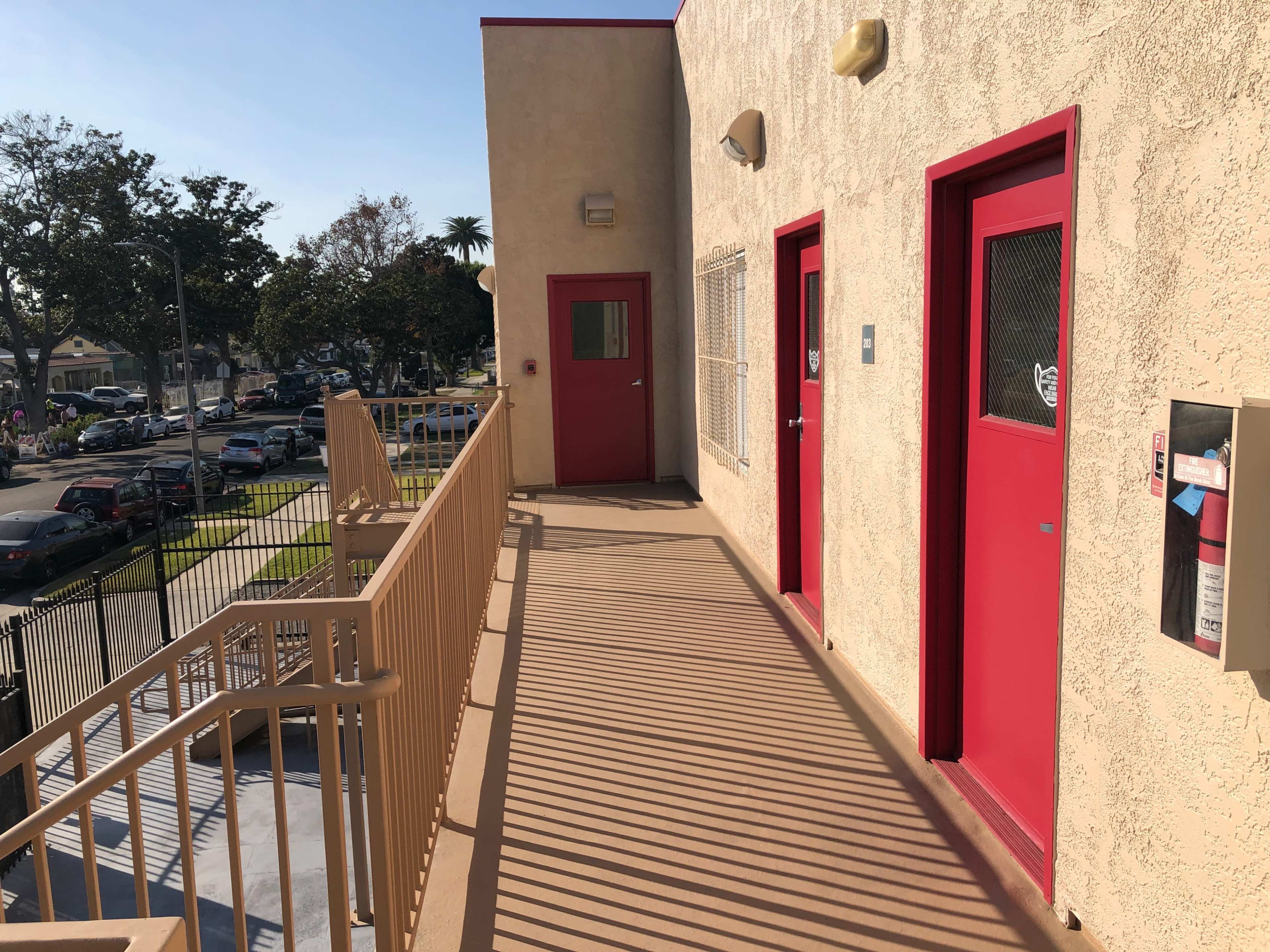 The image shows a sunlit exterior walkway of a building, featuring red doors and a railing, with parked cars visible in the background.
