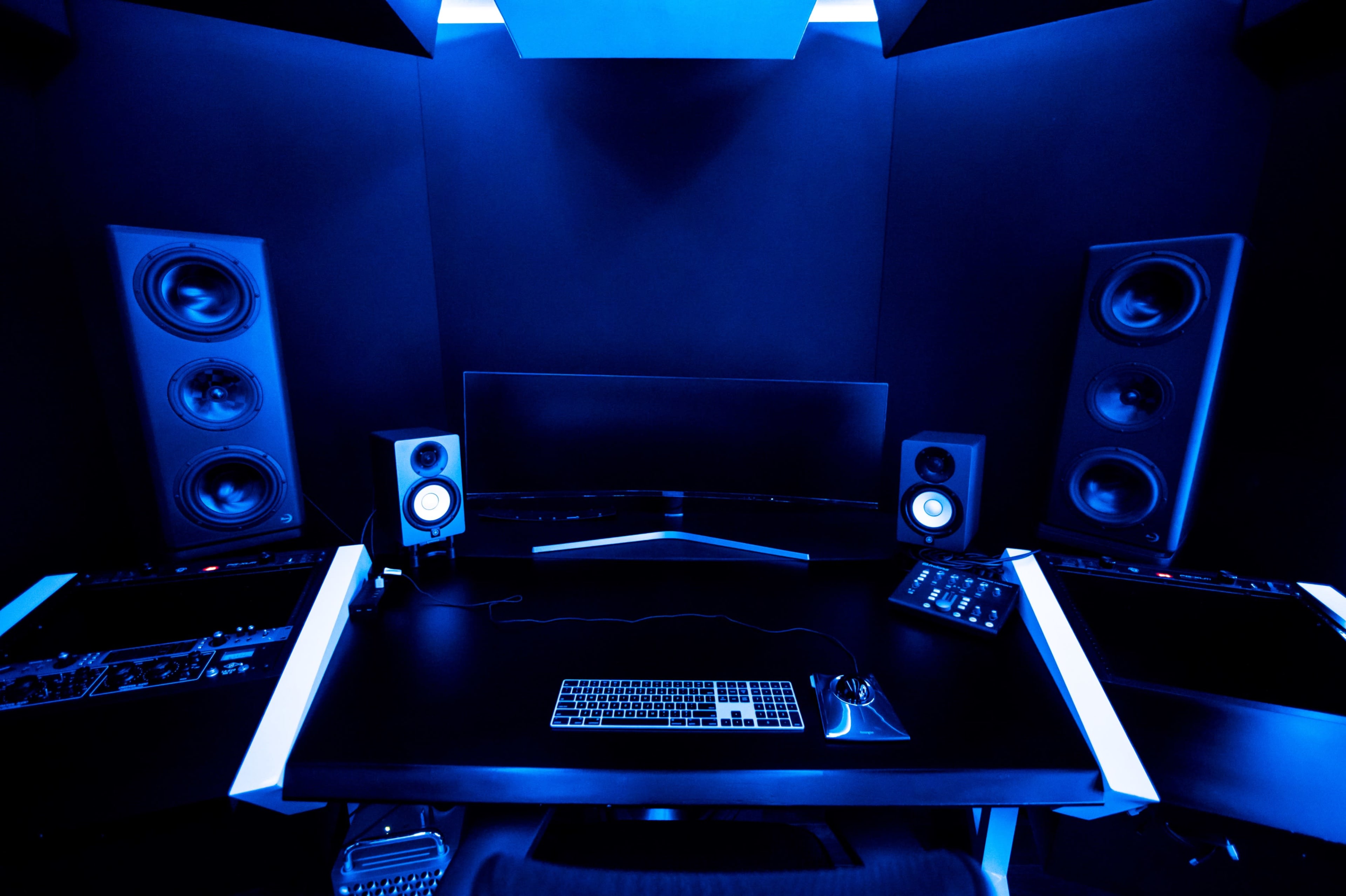 A dark music studio features a desk with a computer monitor, keyboard, and various audio equipment, illuminated by blue lighting.