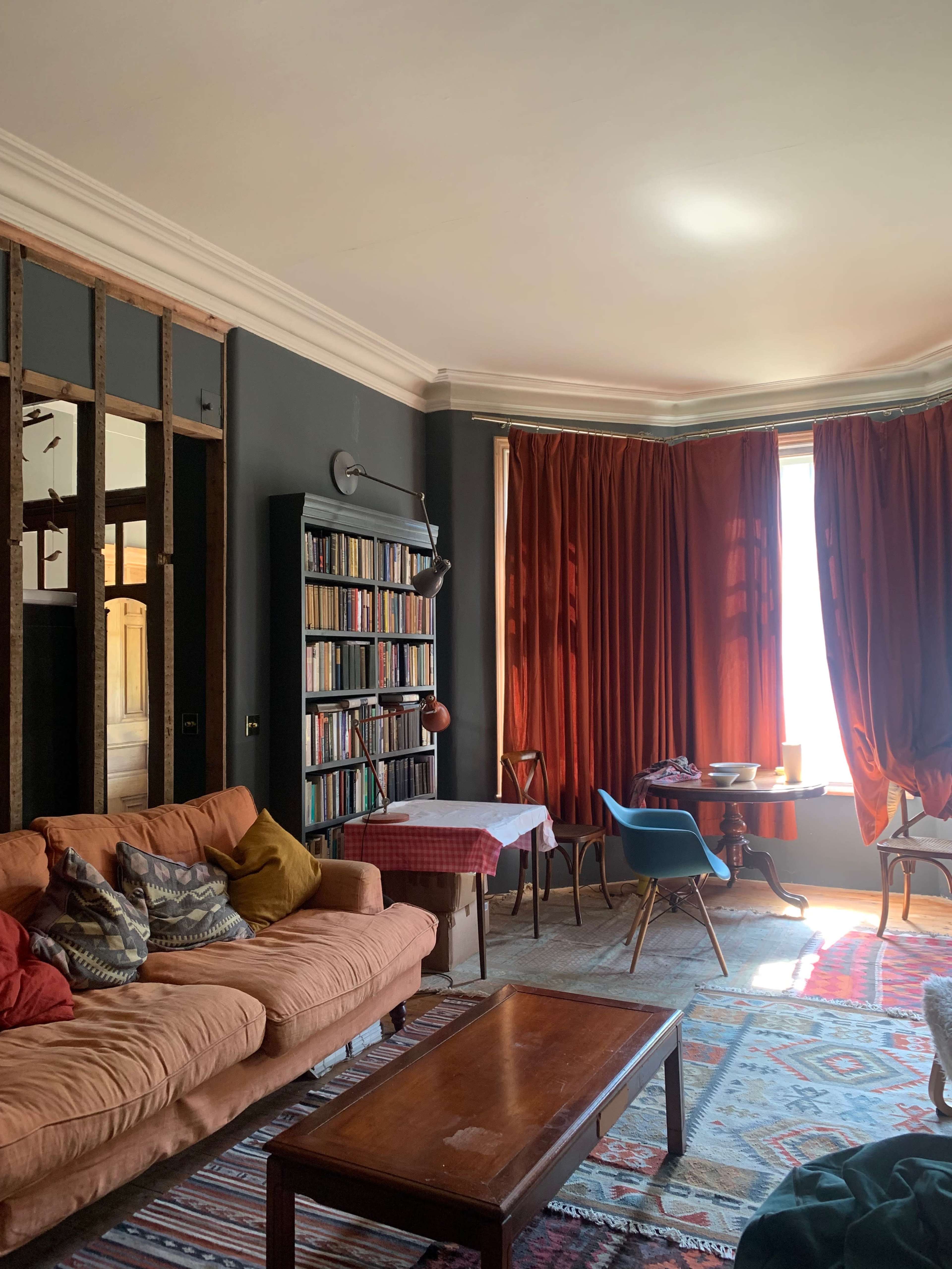 The image shows a cozy living room with a large bookshelf, a terracotta sofa, a wooden coffee table, and a dining area featuring a round table and blue chairs, all illuminated by natural light from tall windows.