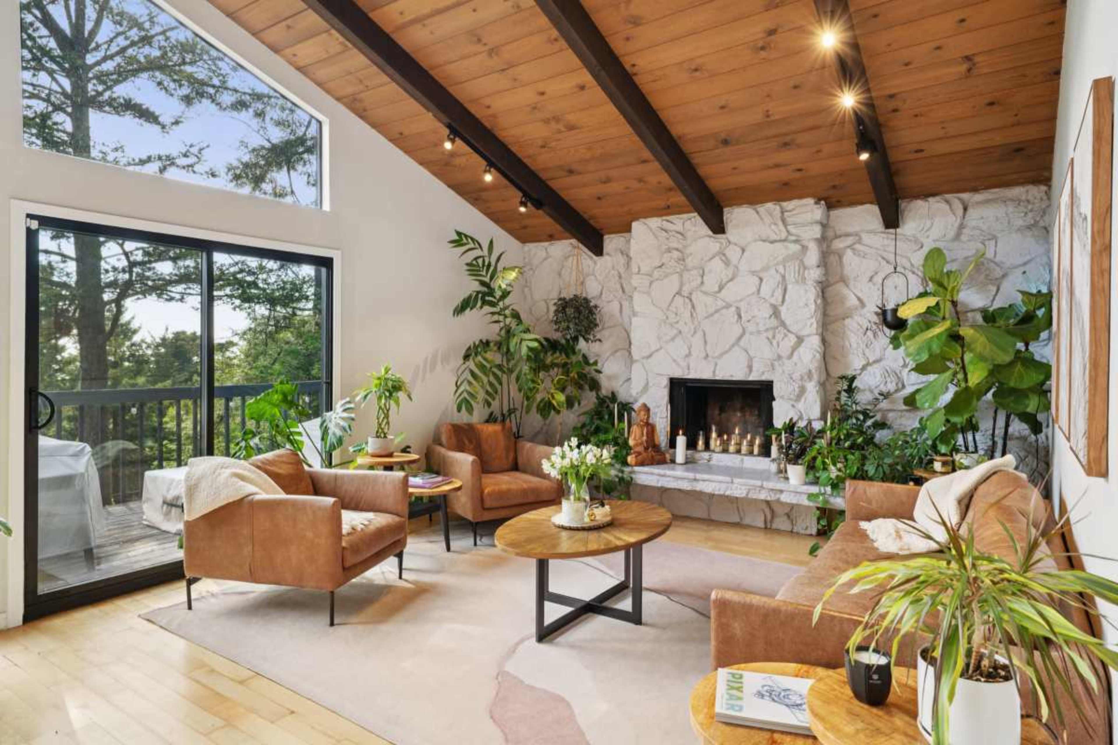 The image shows a spacious living room with a stone fireplace, wooden beams, and several green plants, featuring comfortable leather seating and a coffee table.