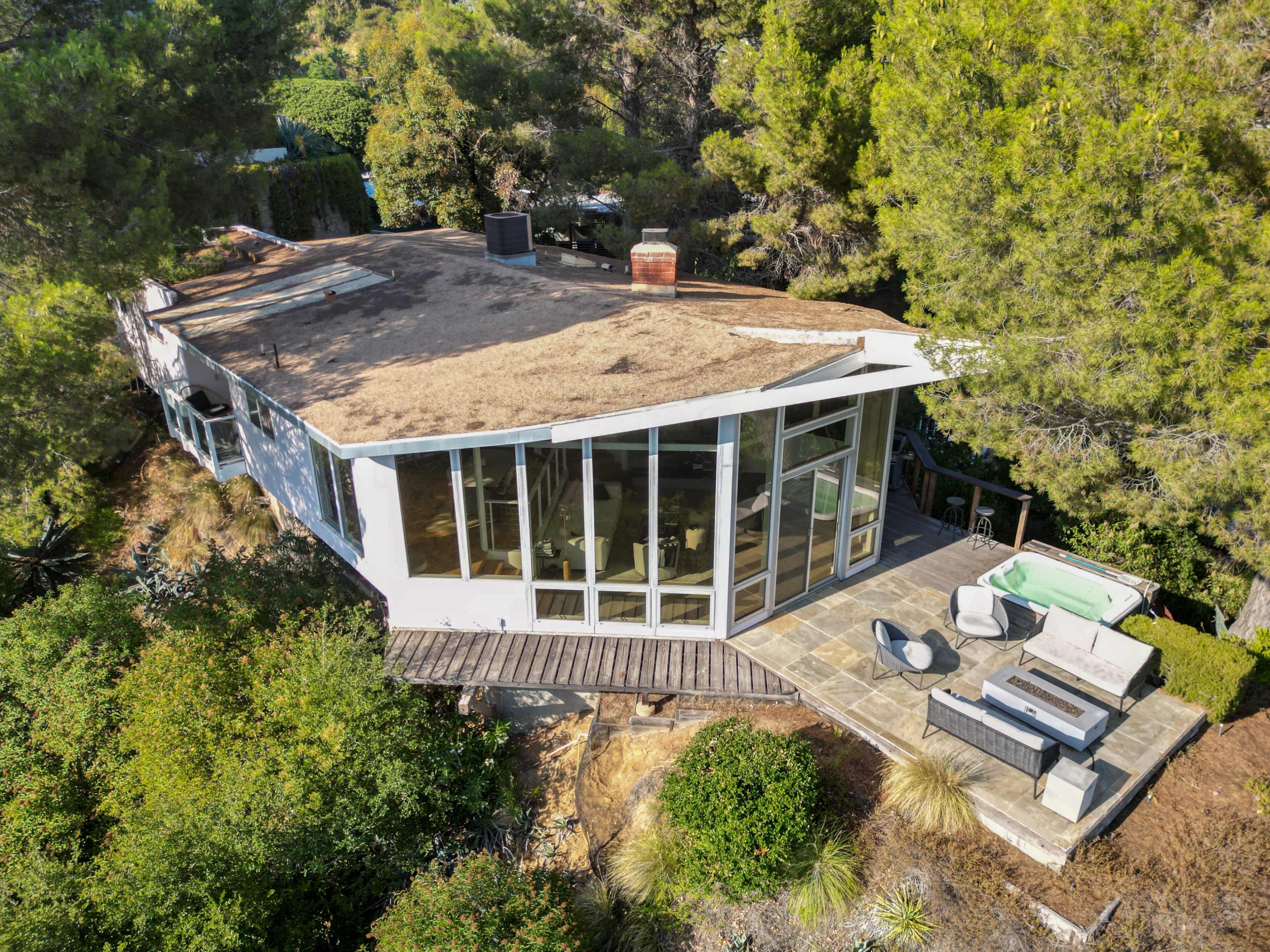 A modern house with a sloped roof is surrounded by greenery and features a stone patio with outdoor seating and a hot tub.
