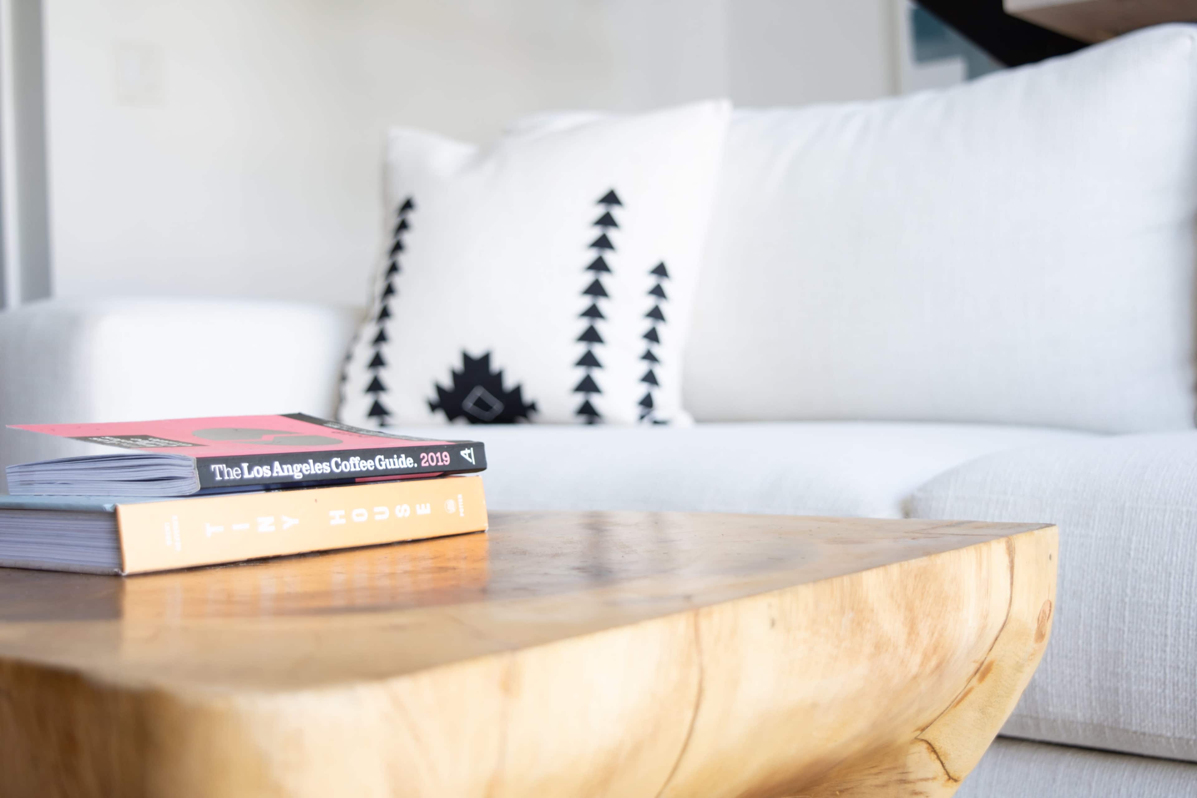 A wooden coffee table holds several books in front of a white couch adorned with a patterned pillow.