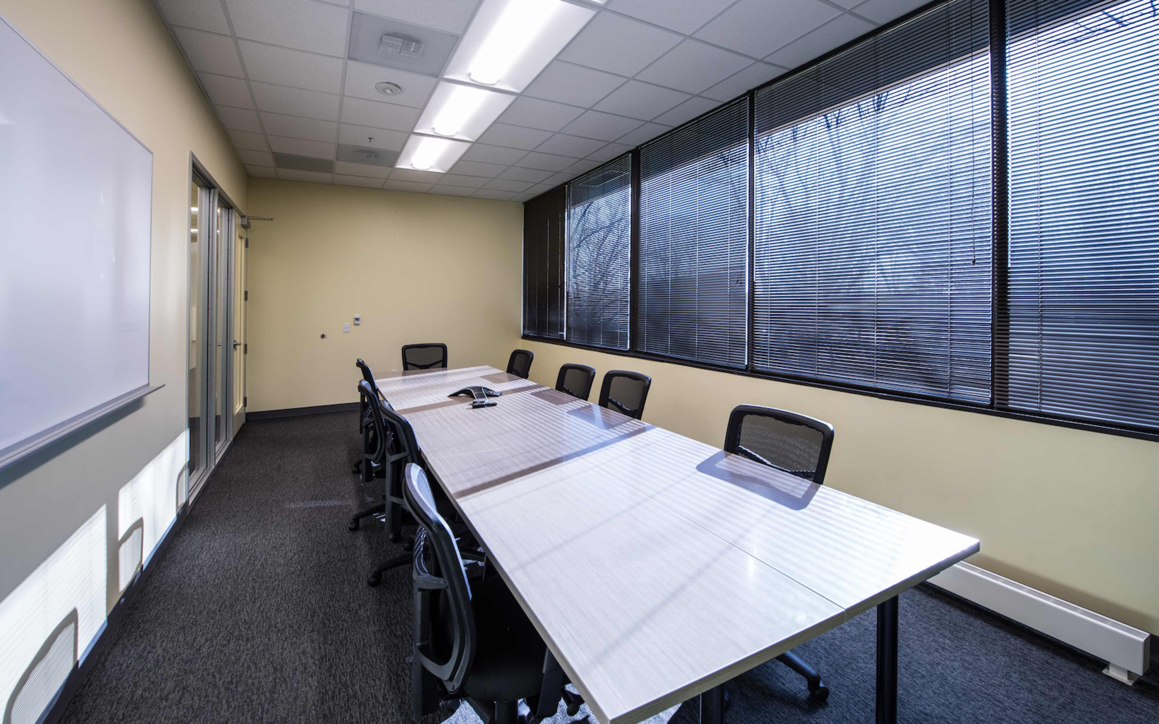A conference room features a long table surrounded by black chairs and large windows with blinds.