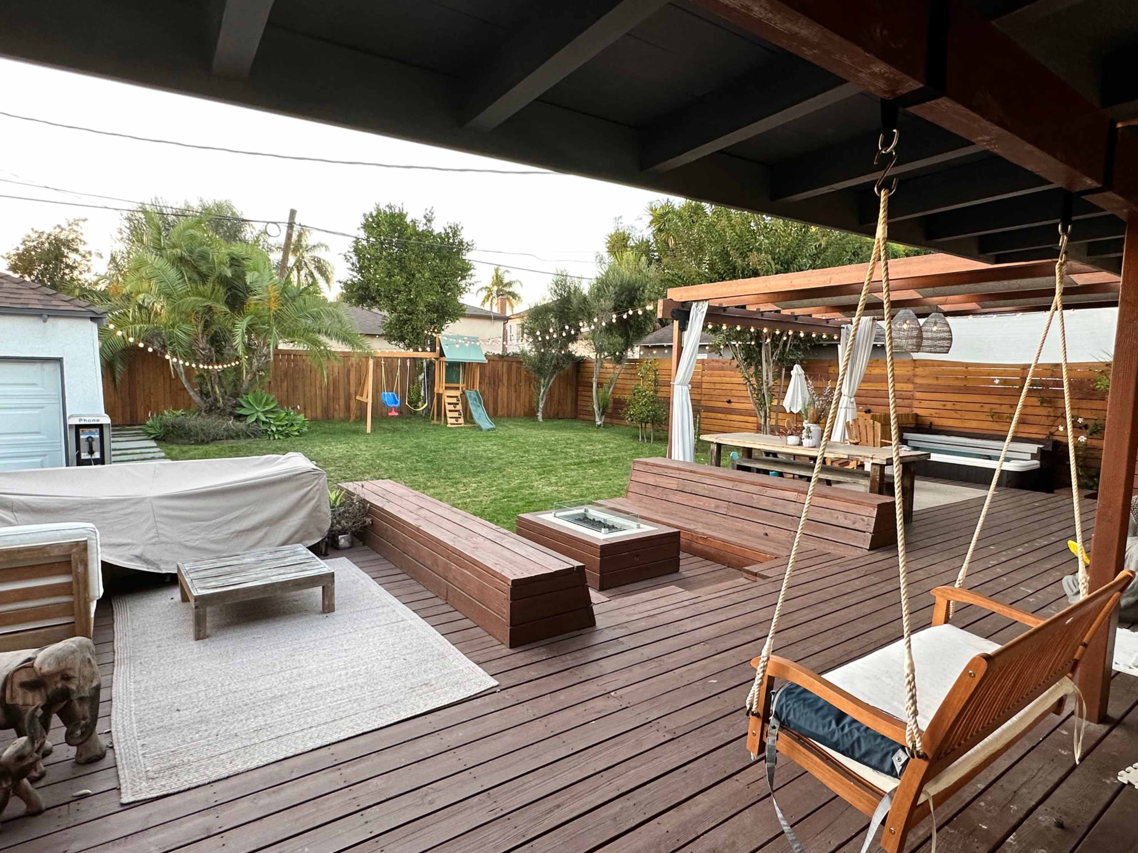 A backyard scene features wooden decking with a seating area, a swing, and a grassy lawn with a playground set in the background.