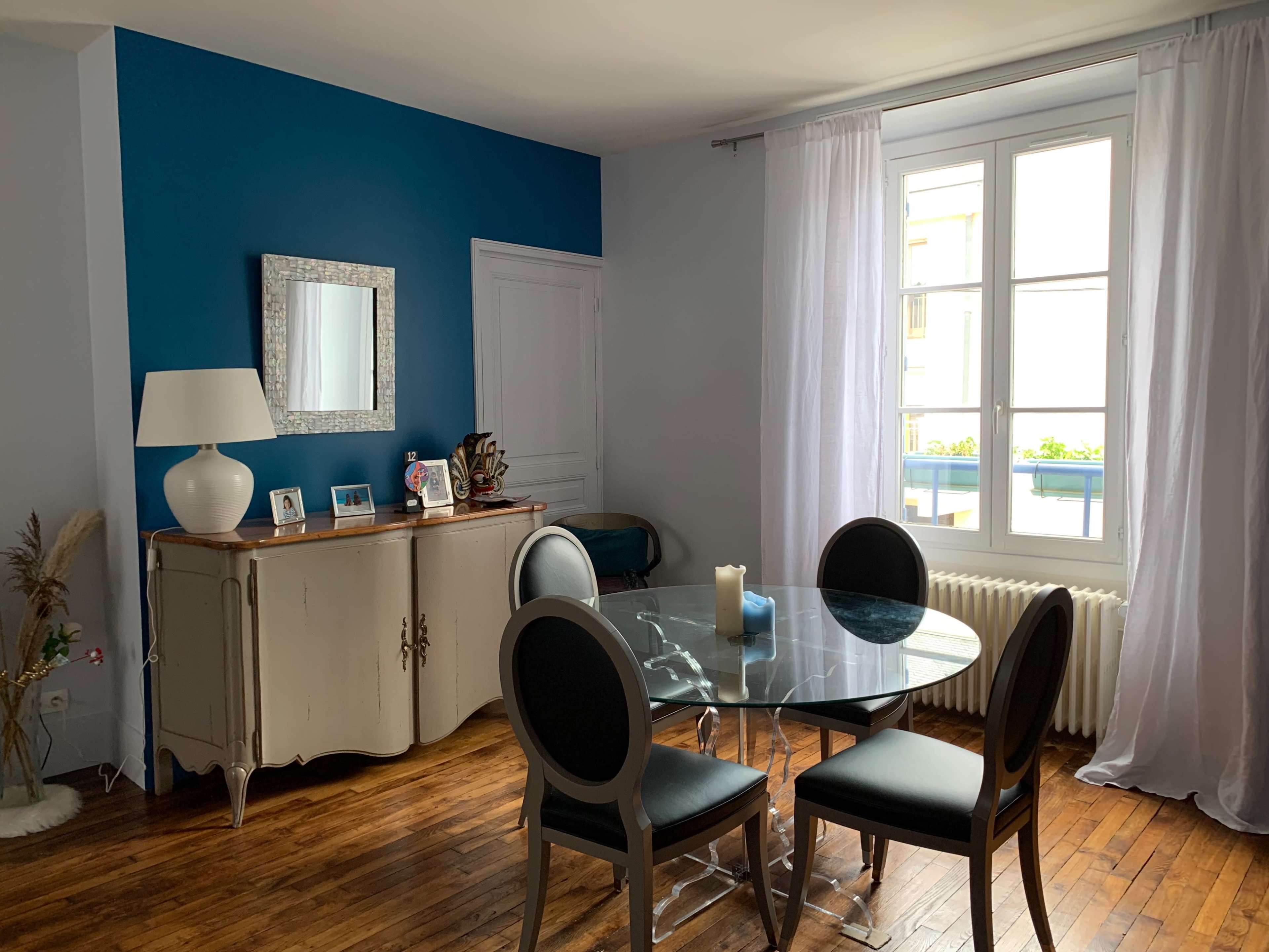 The image shows a dining area with a round glass table surrounded by four black chairs, a sideboard against a blue wall, and a large window covered with white curtains.