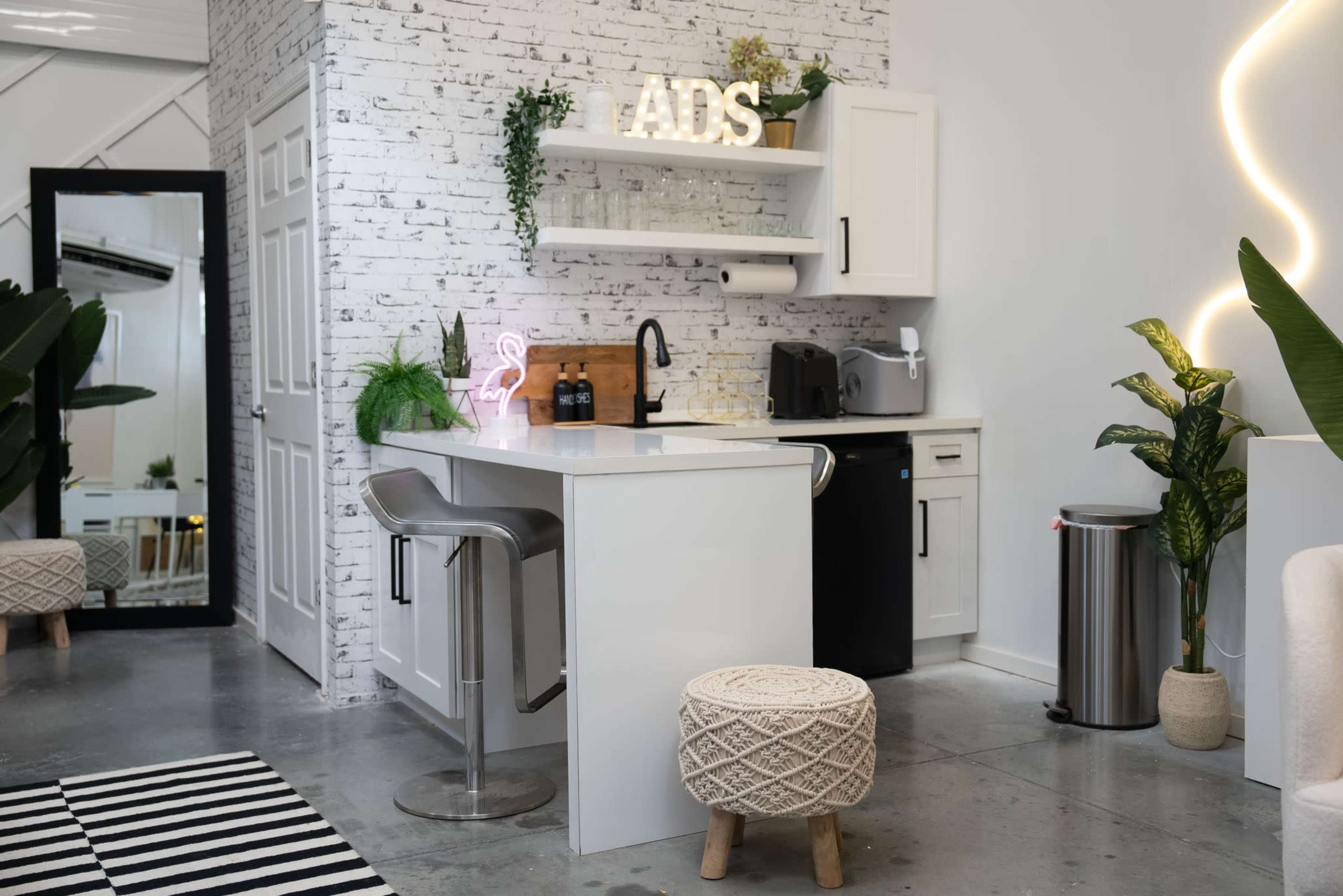 The image shows a modern kitchen area featuring a white bar with stools, a small refrigerator, and decorative plants against a brick wall.