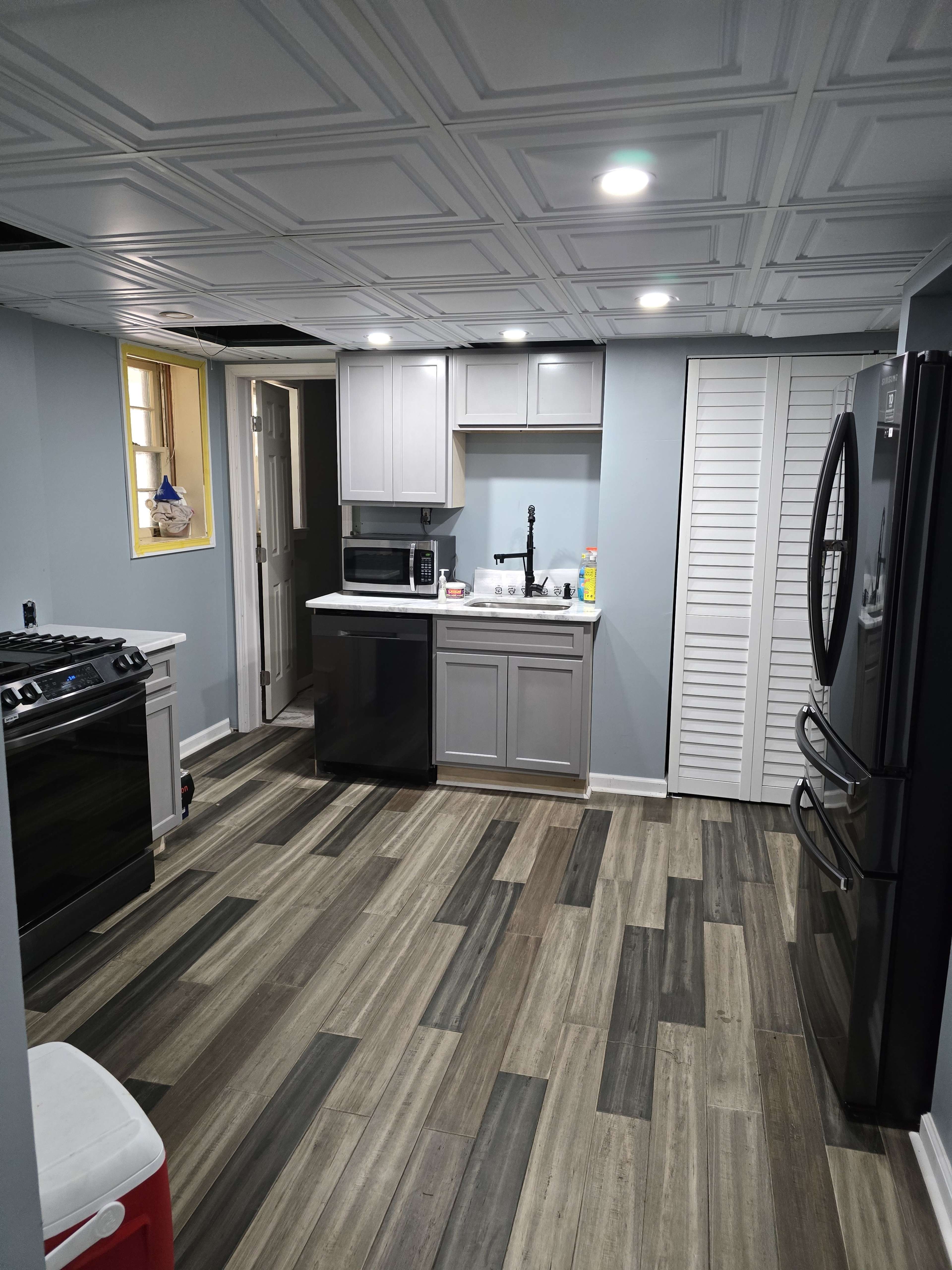 The image shows a modern kitchen with gray cabinets, stainless steel appliances, and a combination of light and dark wood flooring.