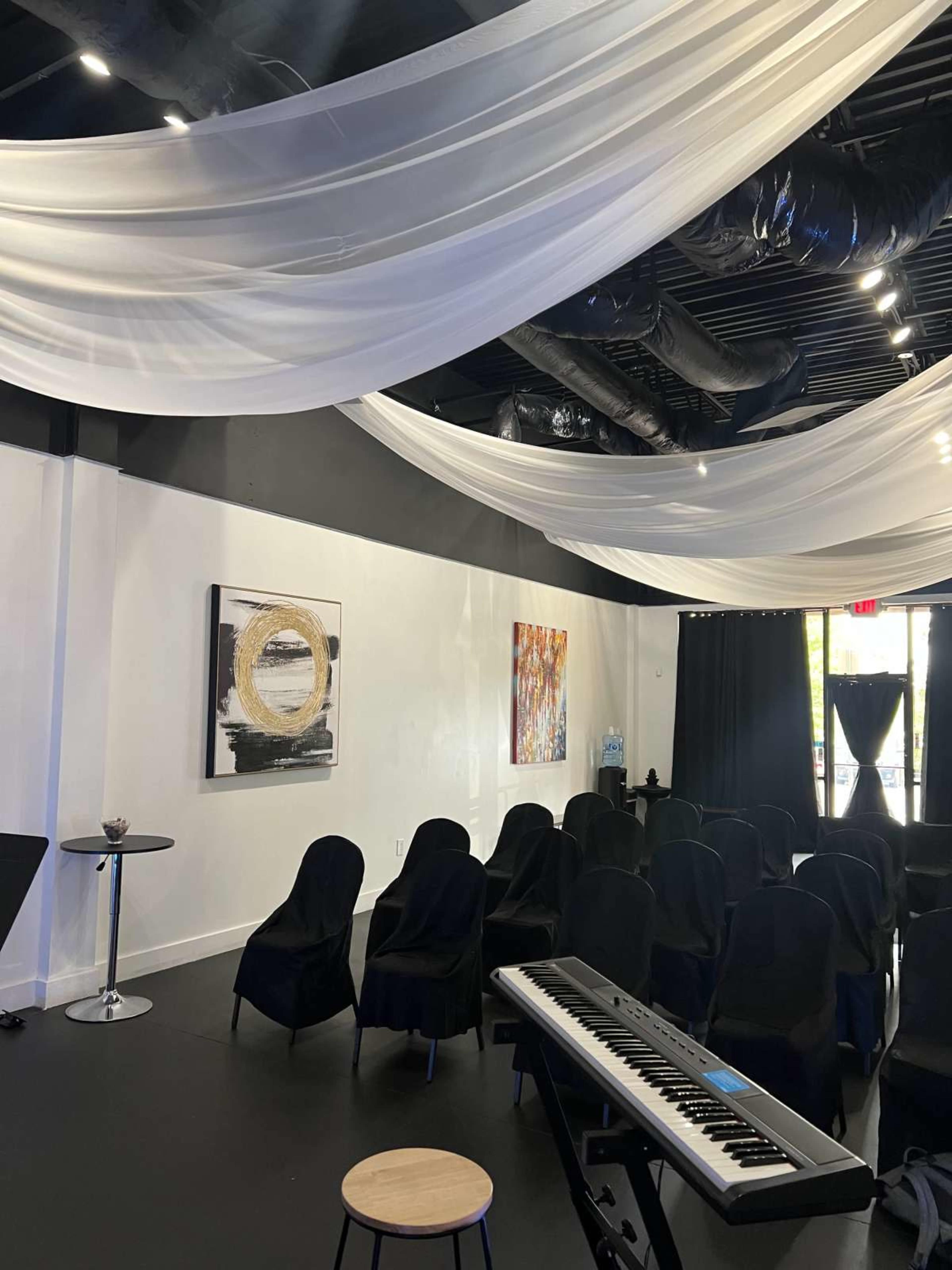 The interior of a venue is set up with black chairs arranged for an event, adorned with white draped fabric overhead, and featuring abstract artwork on the walls.