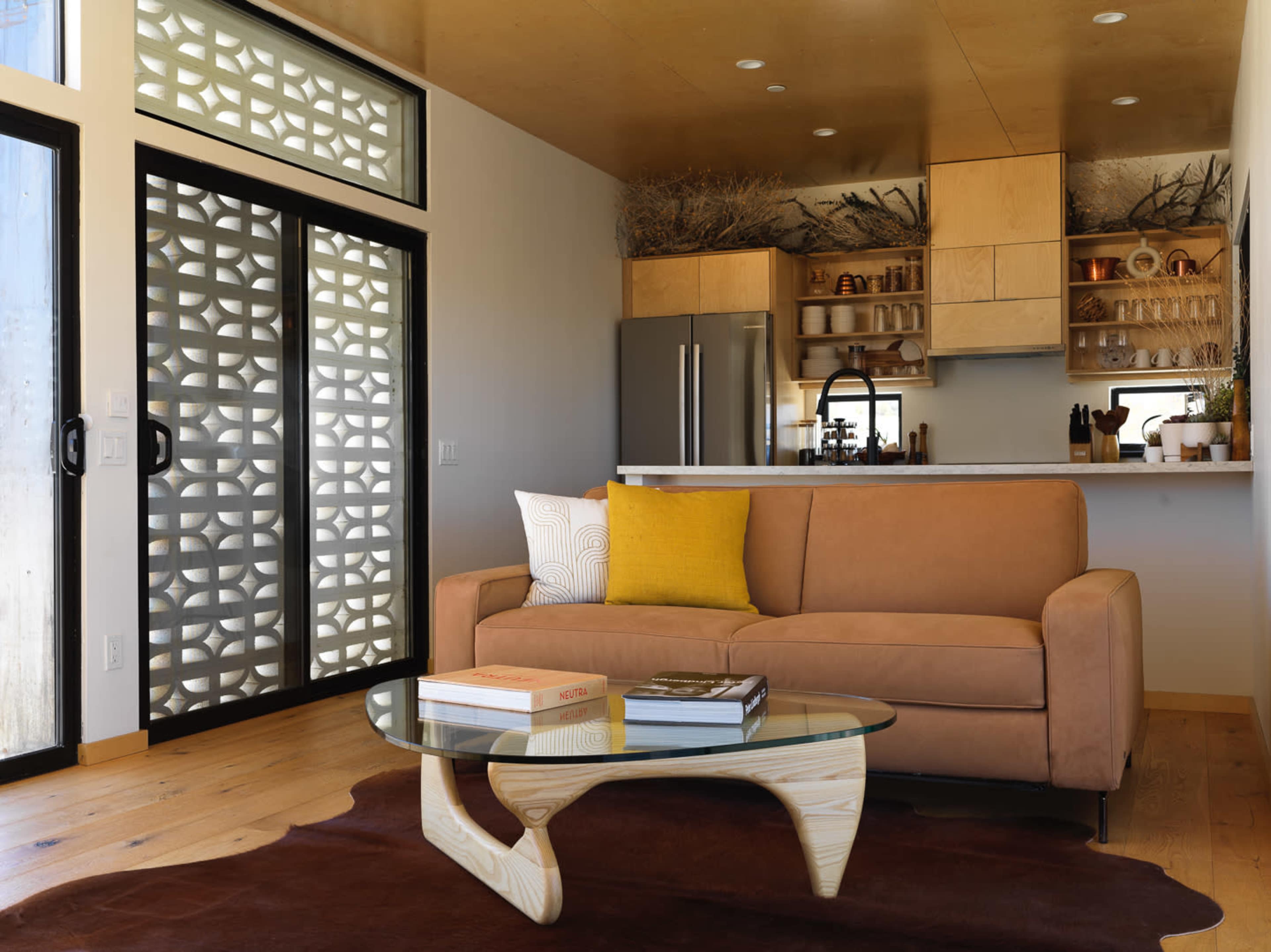 A modern living room featuring a brown sofa, a glass coffee table on a dark rug, and a kitchen area with wooden cabinets in the background.