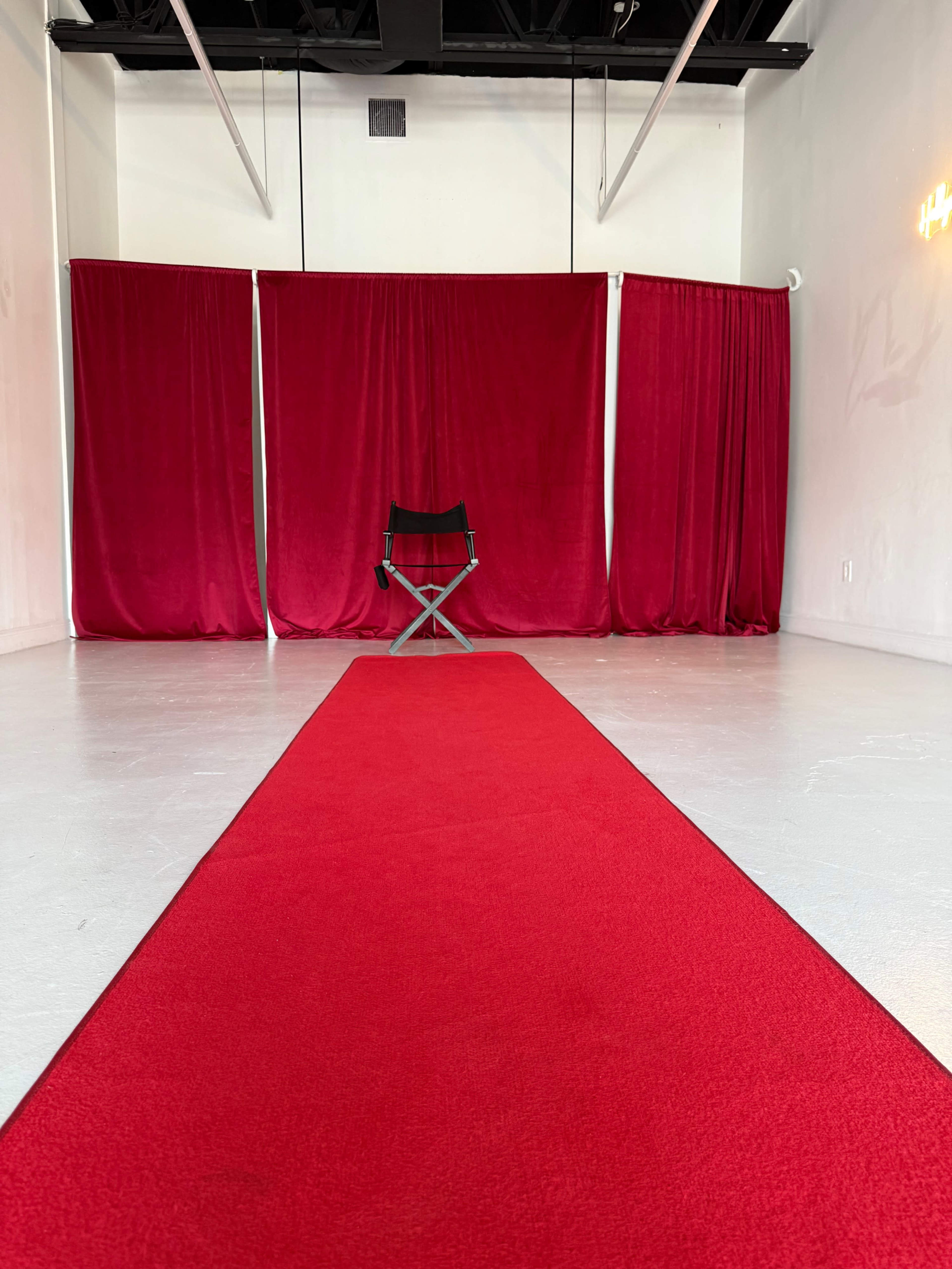 The image shows a red carpet leading to a black director's chair set against a backdrop of red curtains in an empty room.