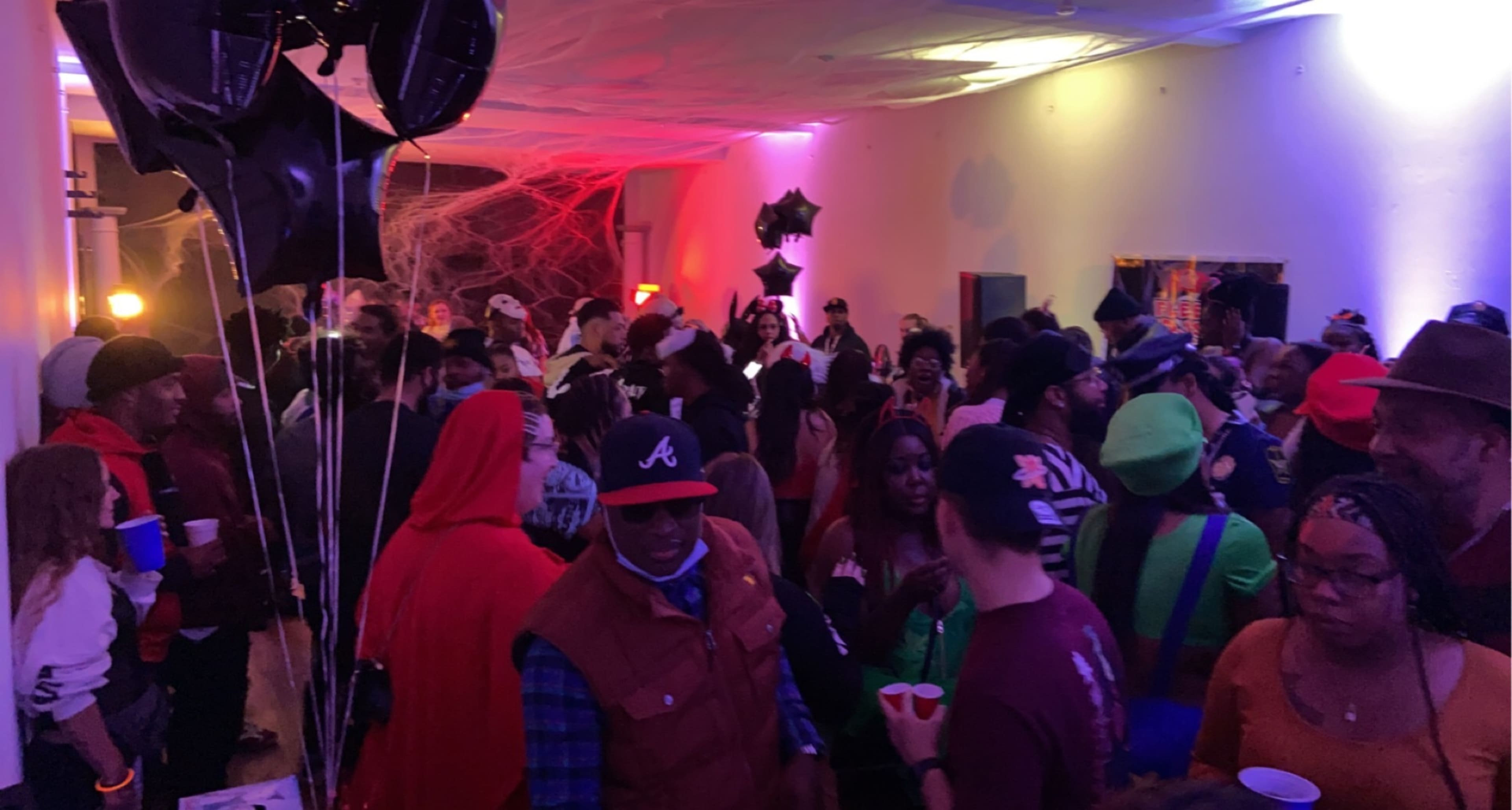A crowded indoor party with people dressed in various costumes, colorful lighting, and decorations visible in the background.