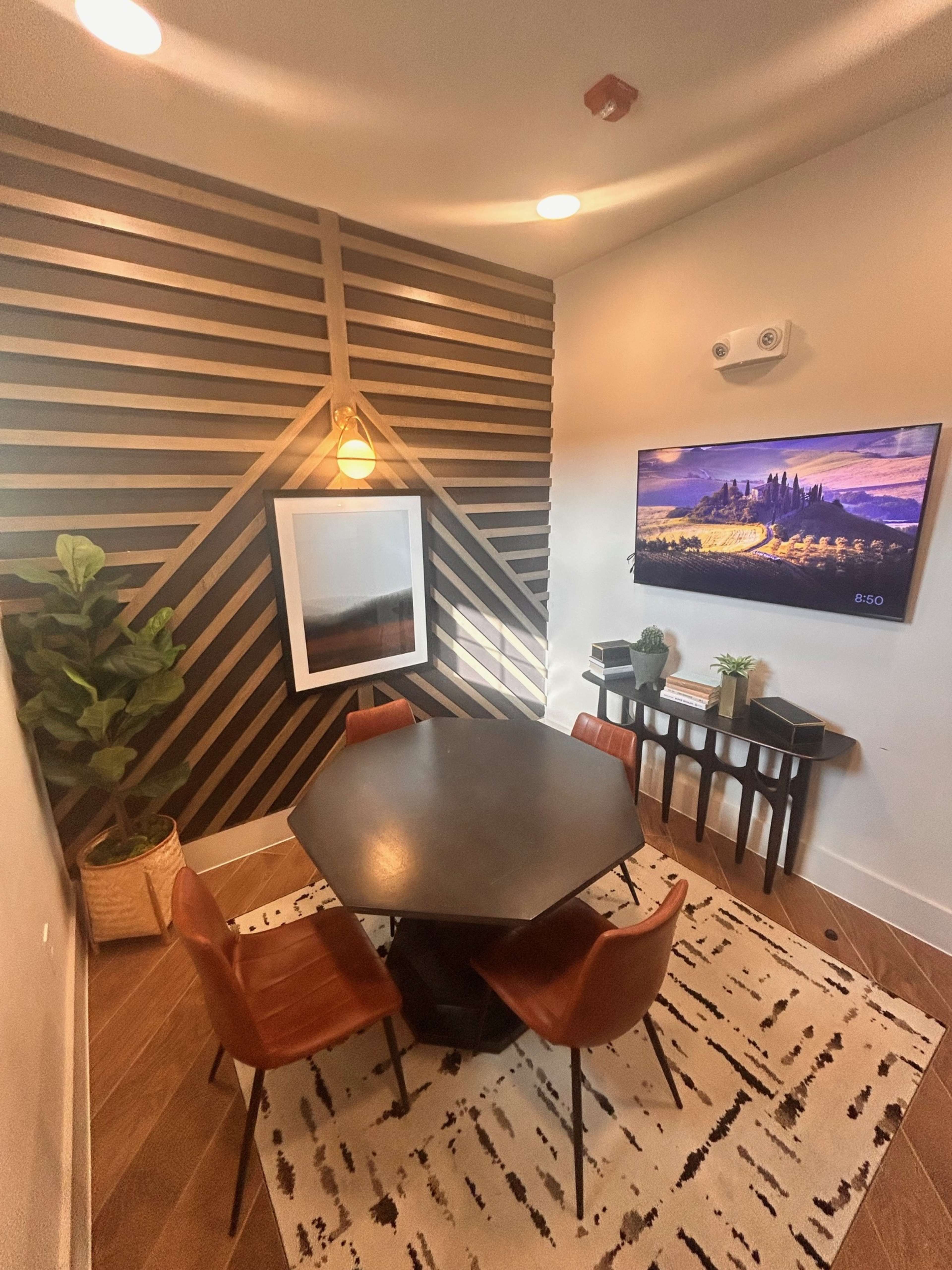 The image shows a small meeting room with a black hexagonal table surrounded by four brown chairs, a large framed picture on the wall, and a television mounted above a slim console table.
