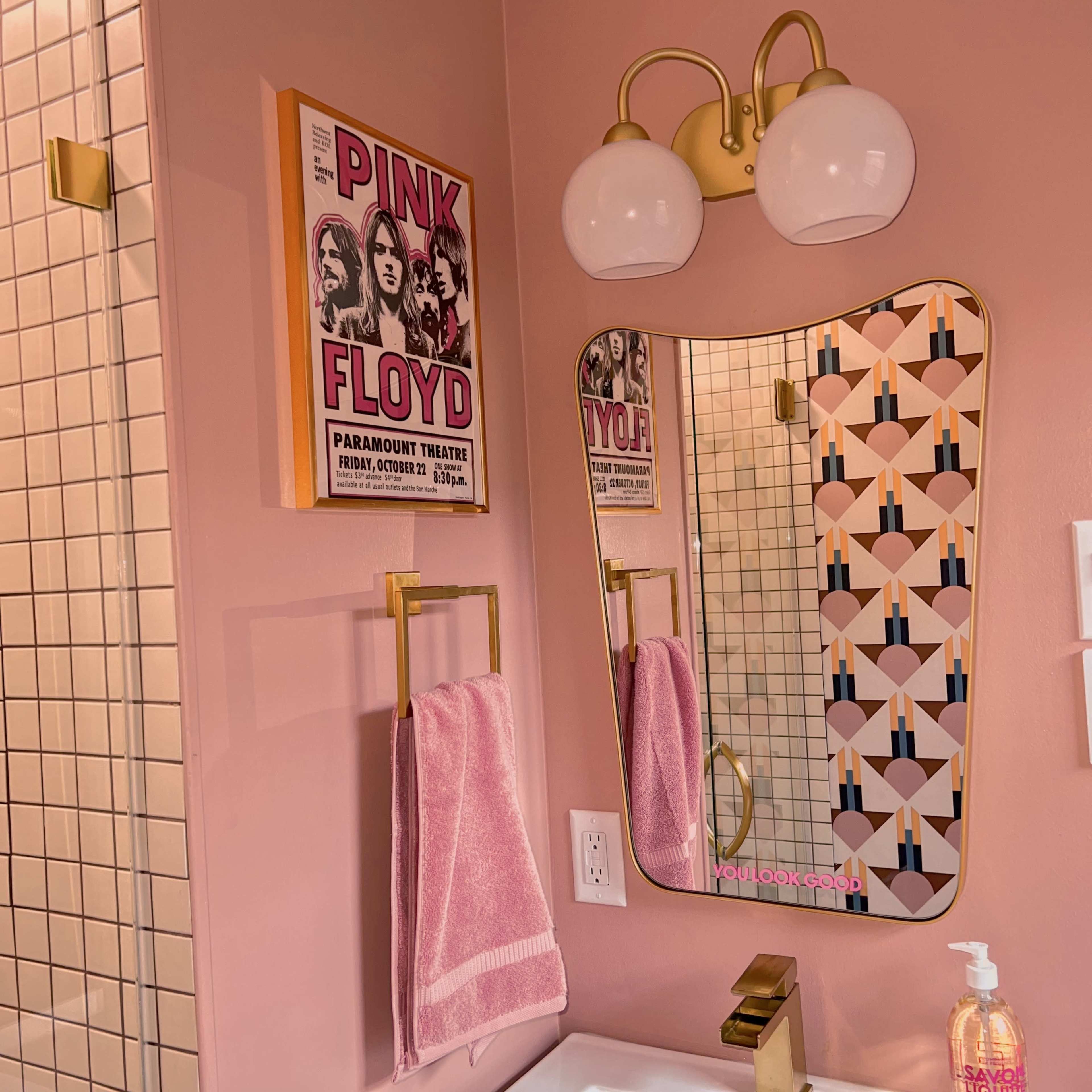 A pink bathroom features a vintage Pink Floyd poster, a stylish mirror with a wavy edge, and a light fixture with two globe bulbs.