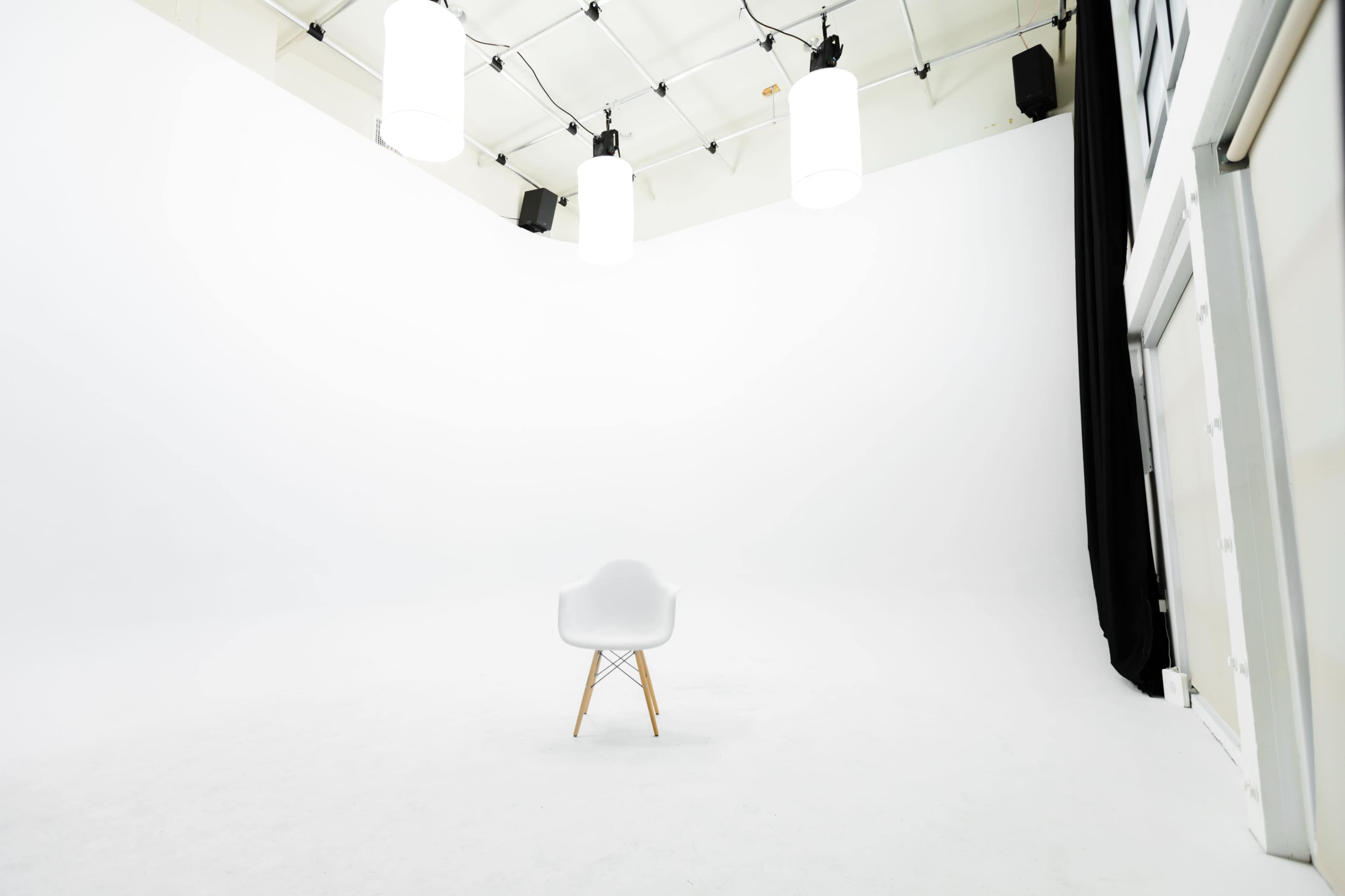 An empty studio space with a single white chair centered on a white floor, illuminated by several hanging lights.