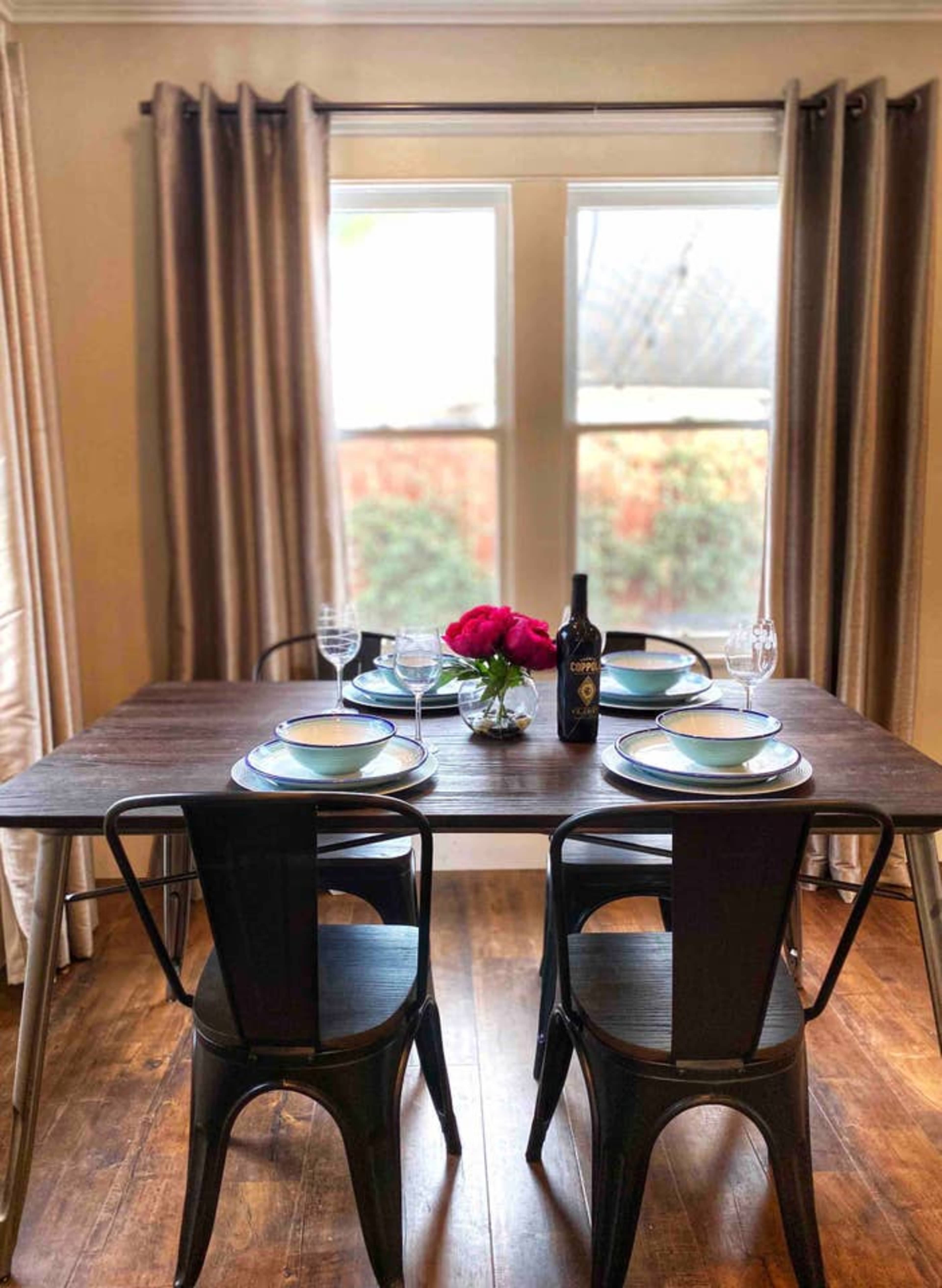 A dining table is set with plates and glasses, featuring a vase of flowers and a bottle of wine, positioned near a window with curtains.