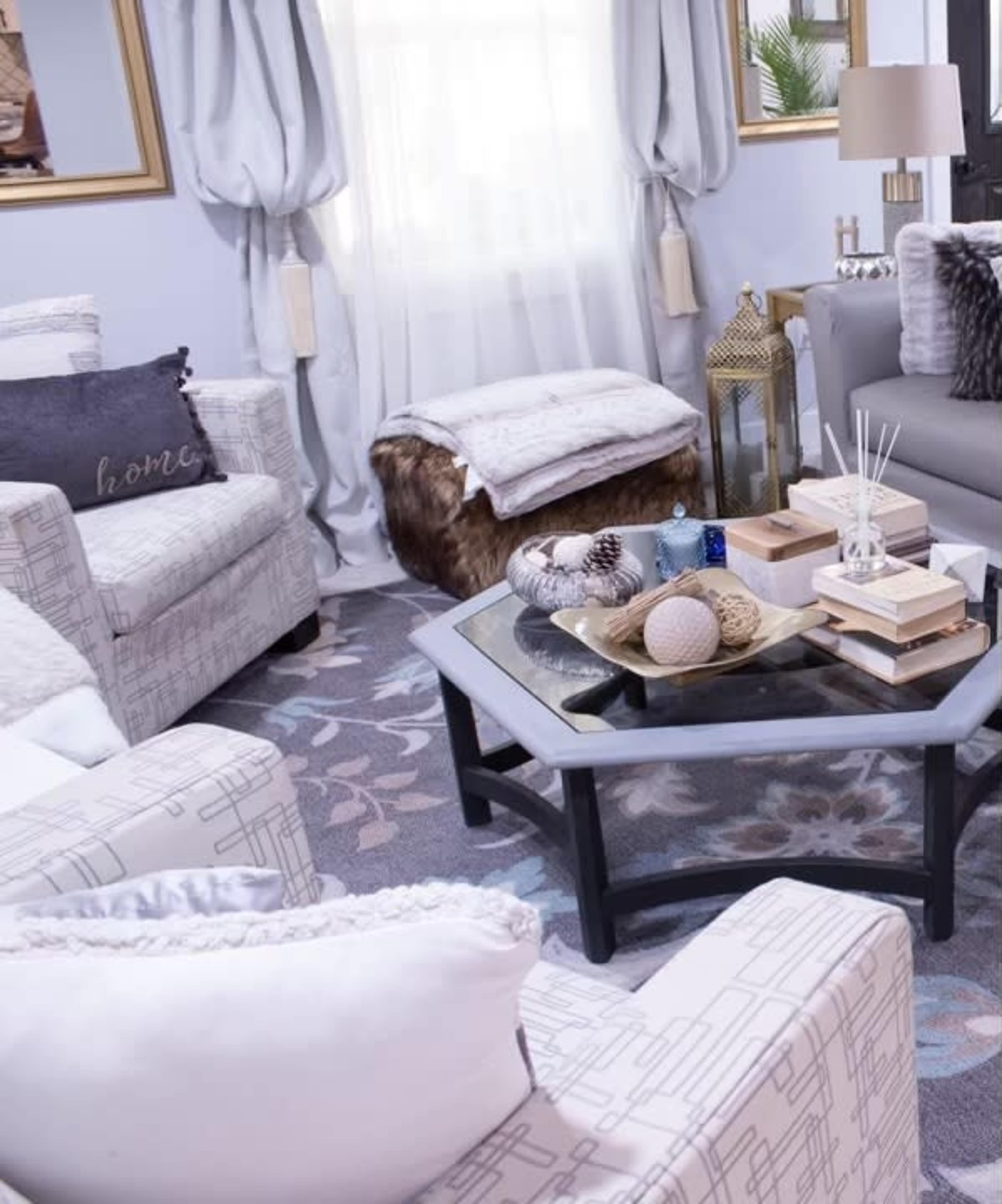 The image shows a cozy living room with two patterned armchairs, a coffee table adorned with decorative items, and large windows draped with curtains, creating a warm atmosphere.