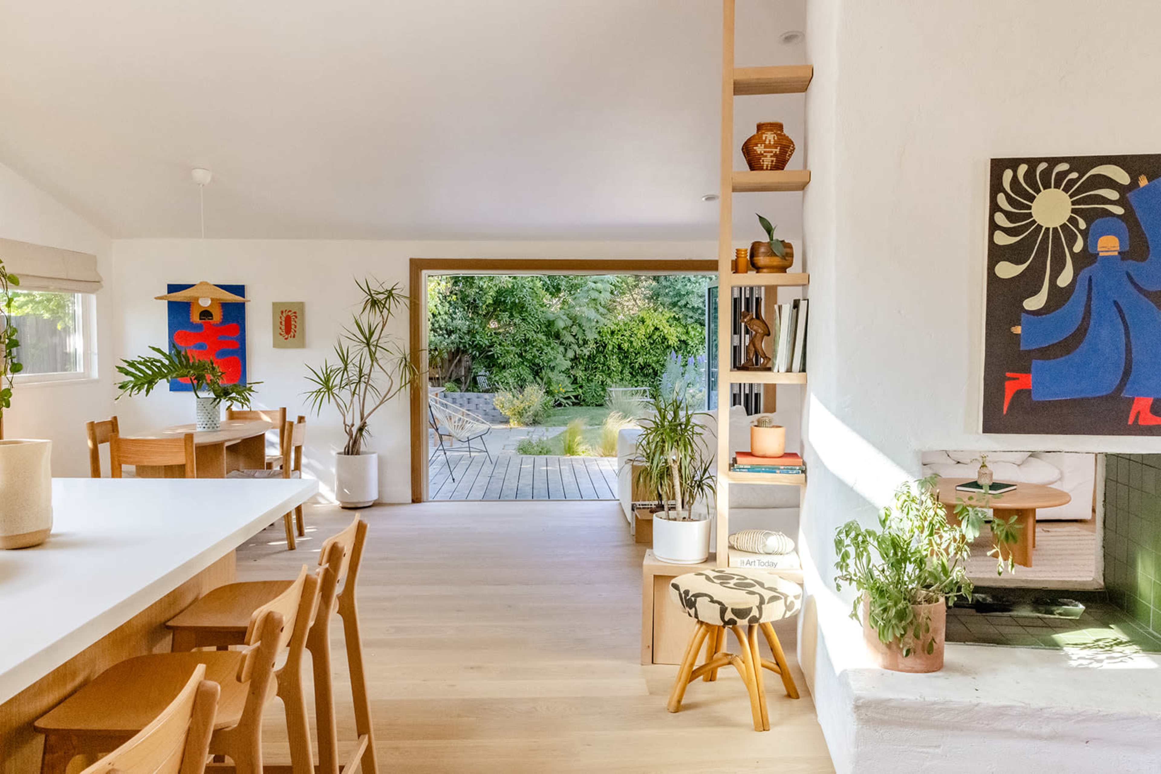 A modern living space features a dining area with wooden chairs and a large table, opening up to a garden through a sliding glass door.