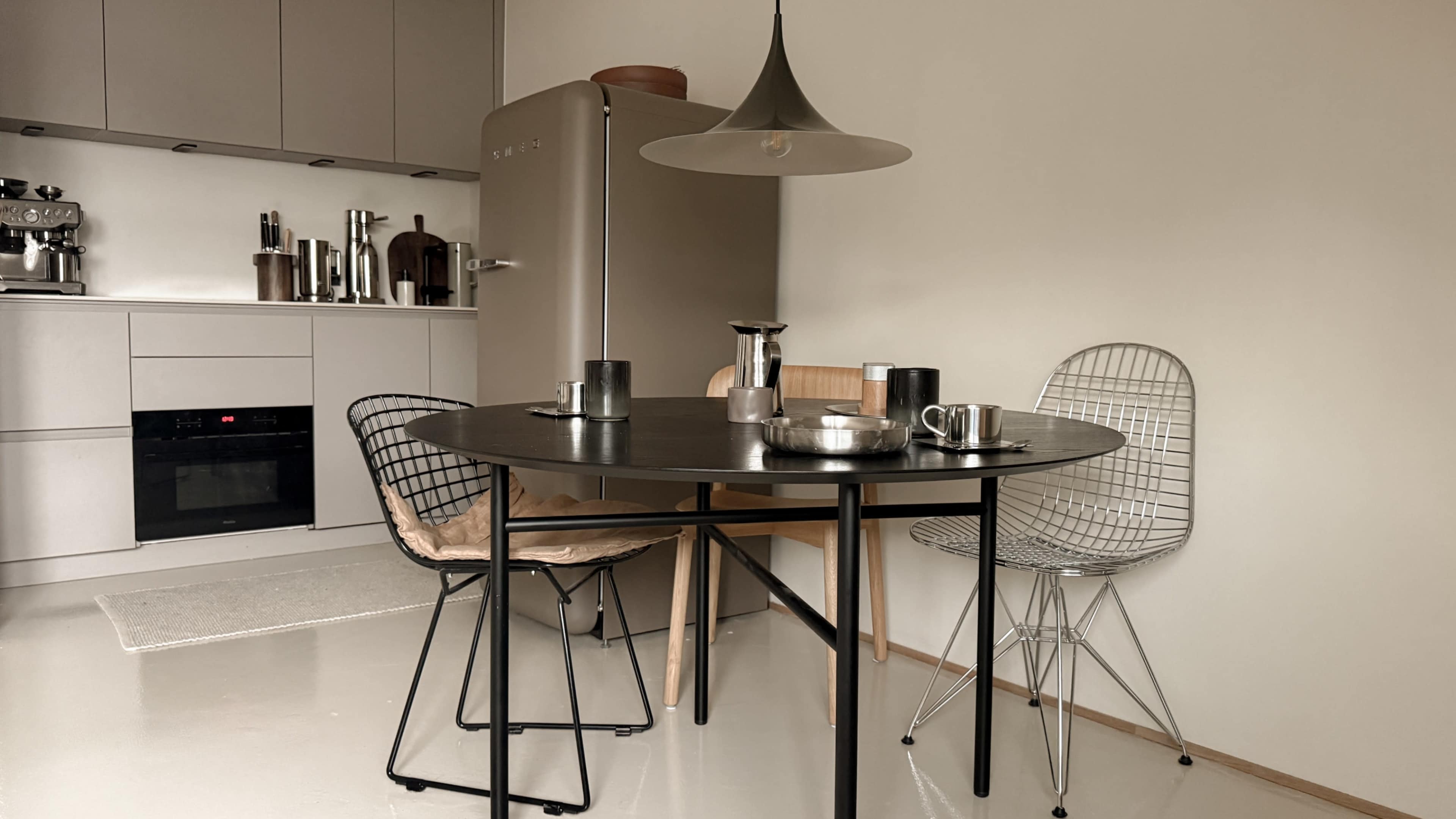 A dining area features a round black table with various metal serving pieces and three different chairs, set against a backdrop of a modern kitchen with gray cabinets and a stainless steel refrigerator.