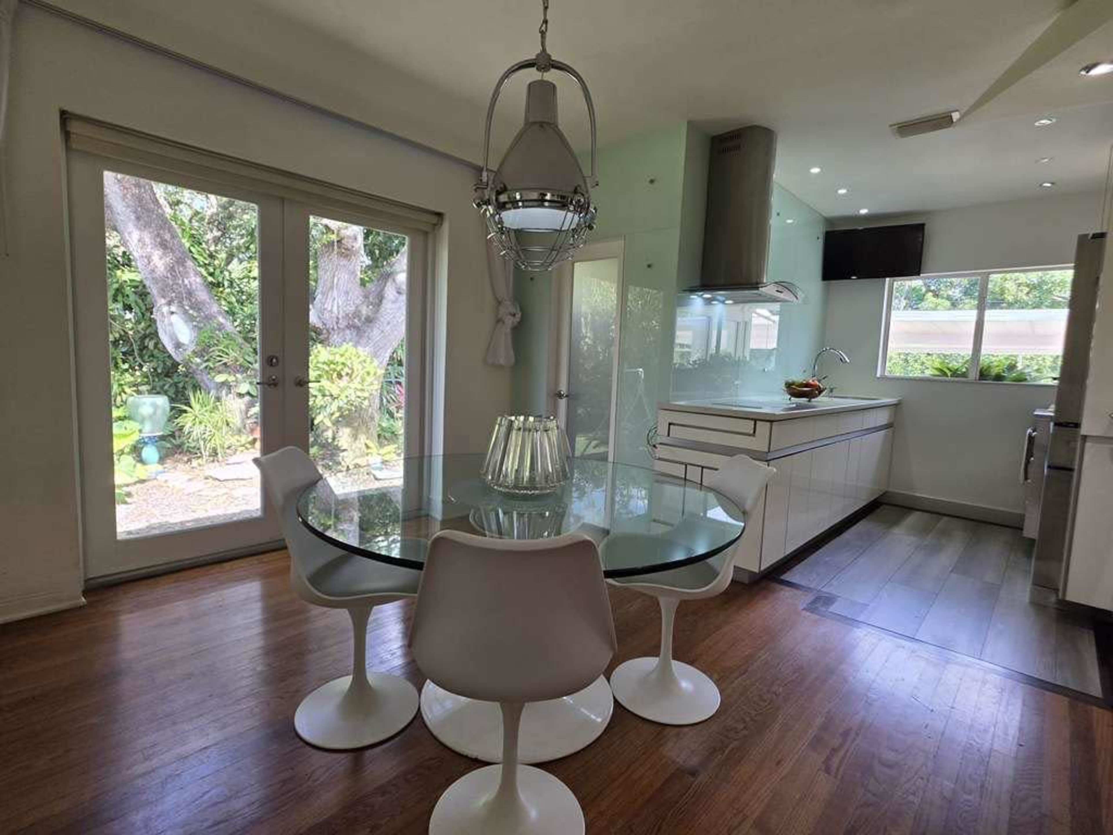 The image shows a modern dining area with a round glass table and four white chairs, adjacent to a kitchen with sleek cabinetry and a view of a garden through large windows.