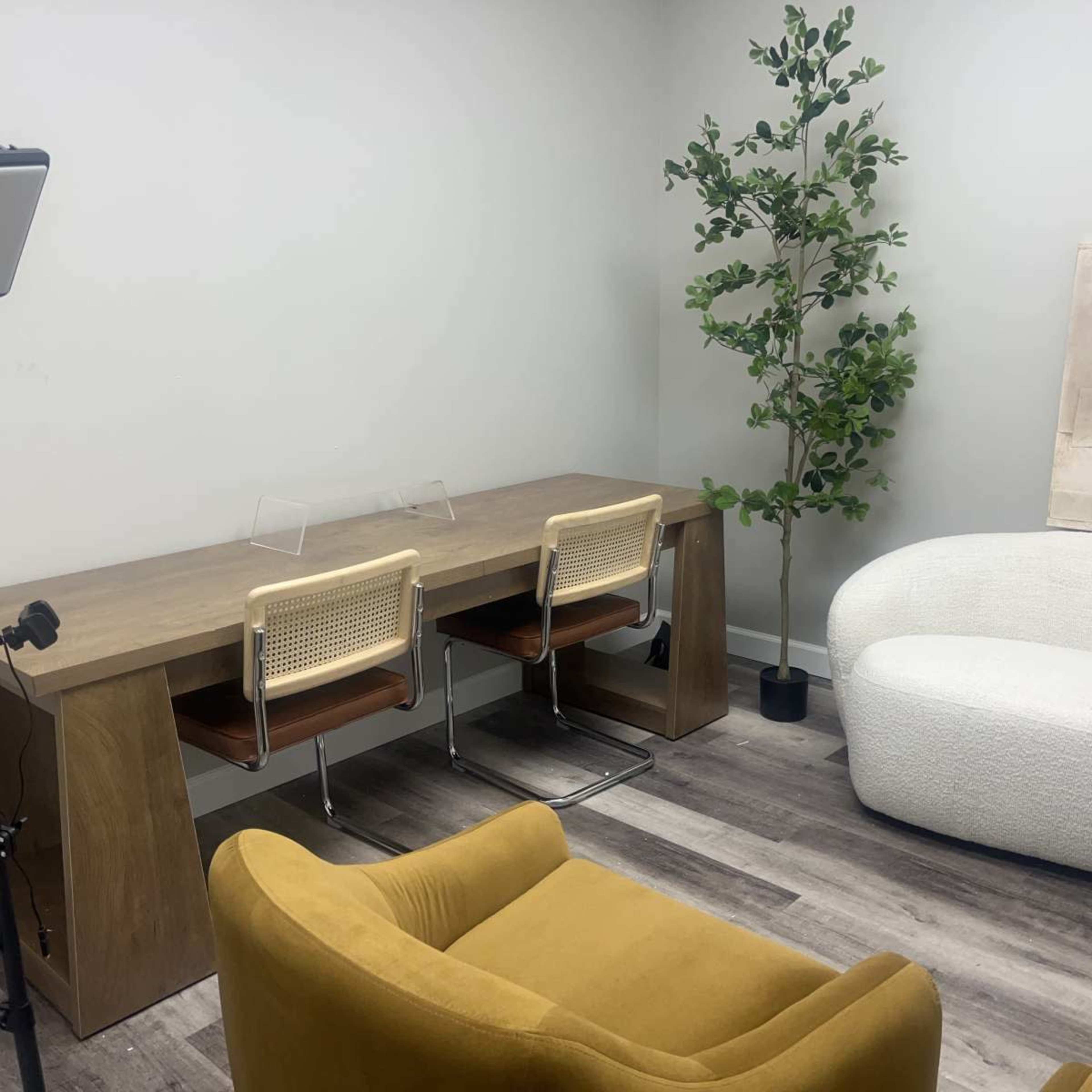 The image shows a minimalist office space featuring a wooden desk with two chairs, a small decorative plant, and a curved white sofa.