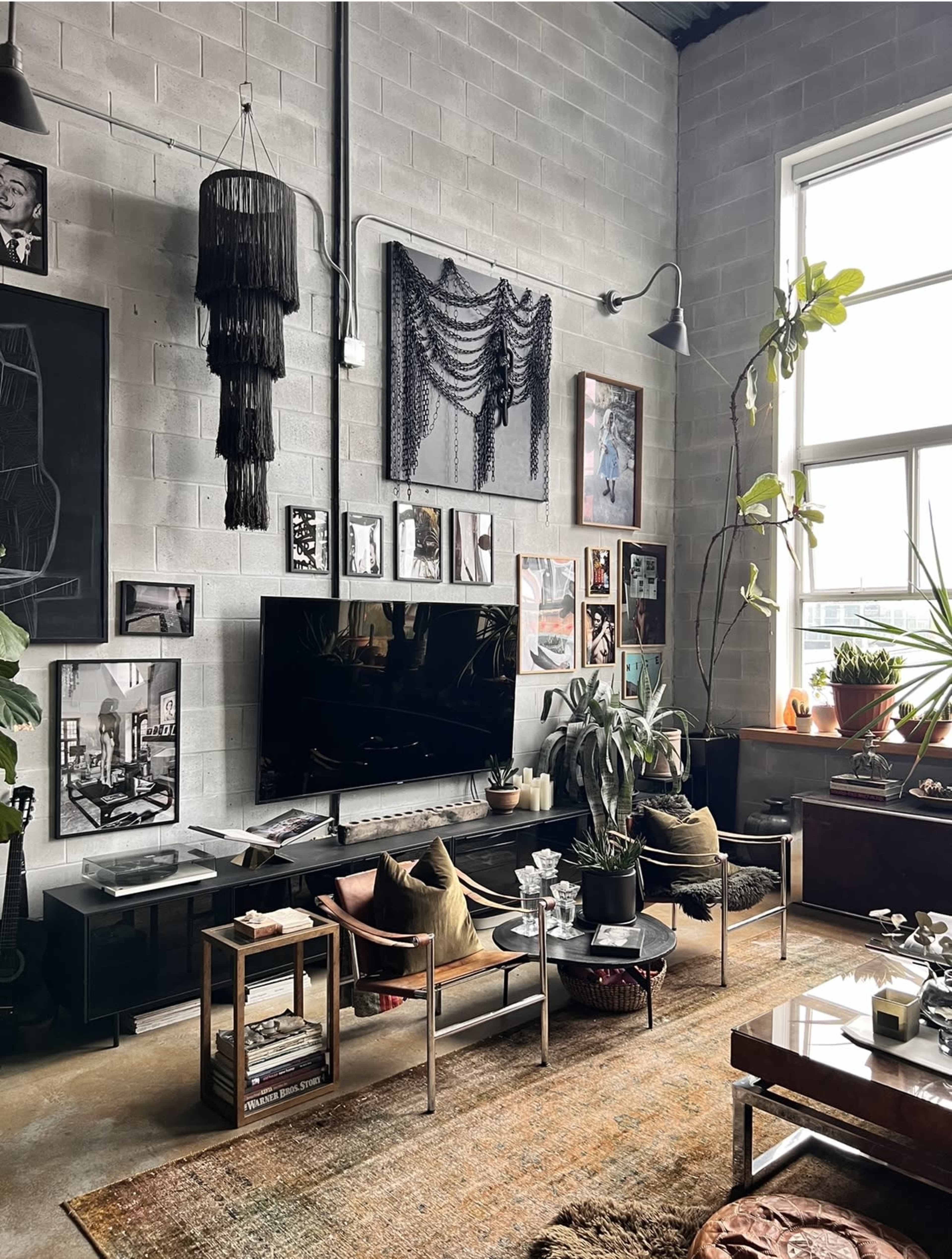 A modern living room features a large television, a variety of wall art, potted plants, and a textured rug, with industrial-style decor elements and ample natural light from a large window.