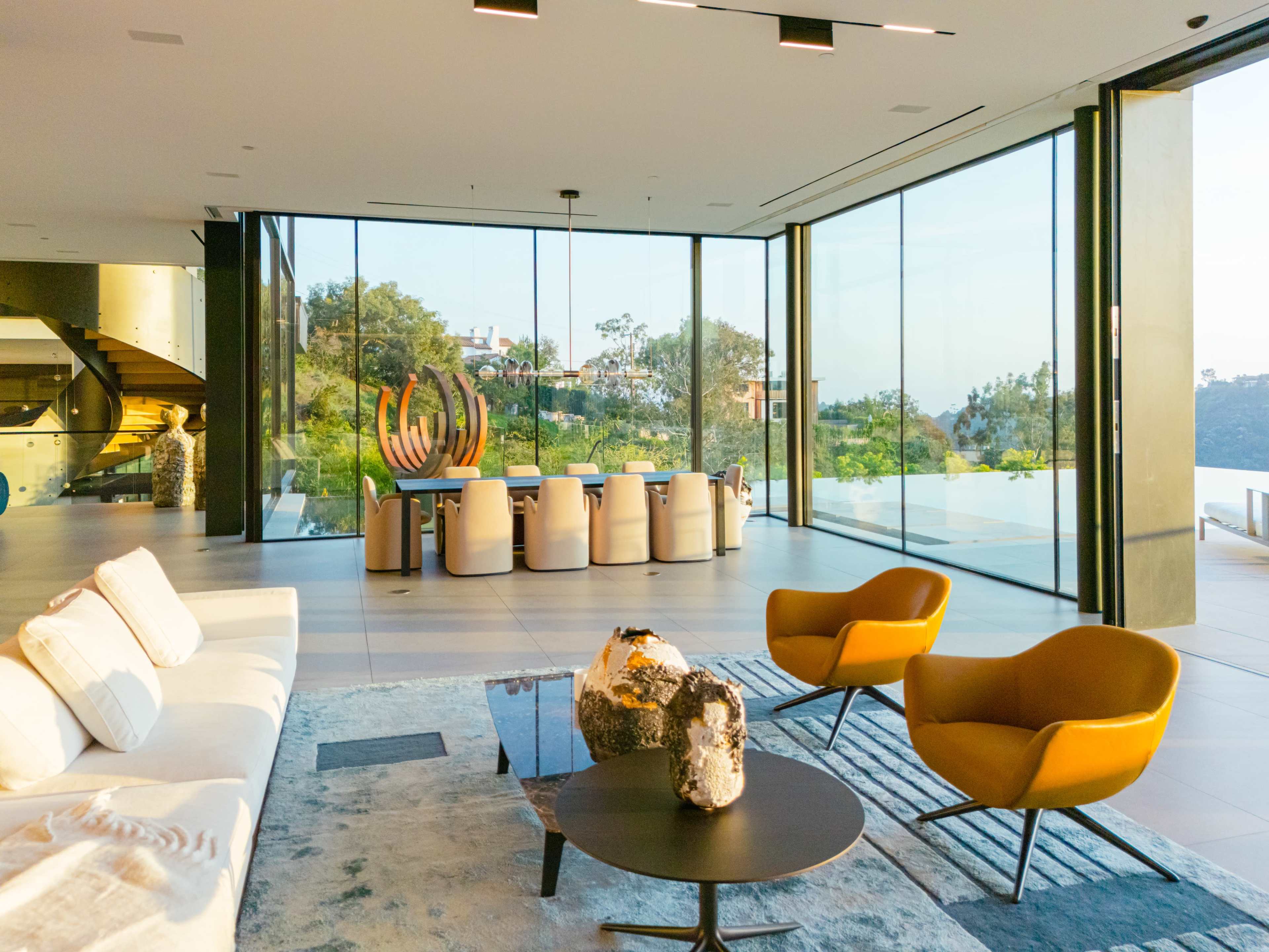 A modern living area with large glass walls, featuring a comfortable sofa, two orange chairs, a black coffee table, and a dining area overlooking a landscaped exterior.