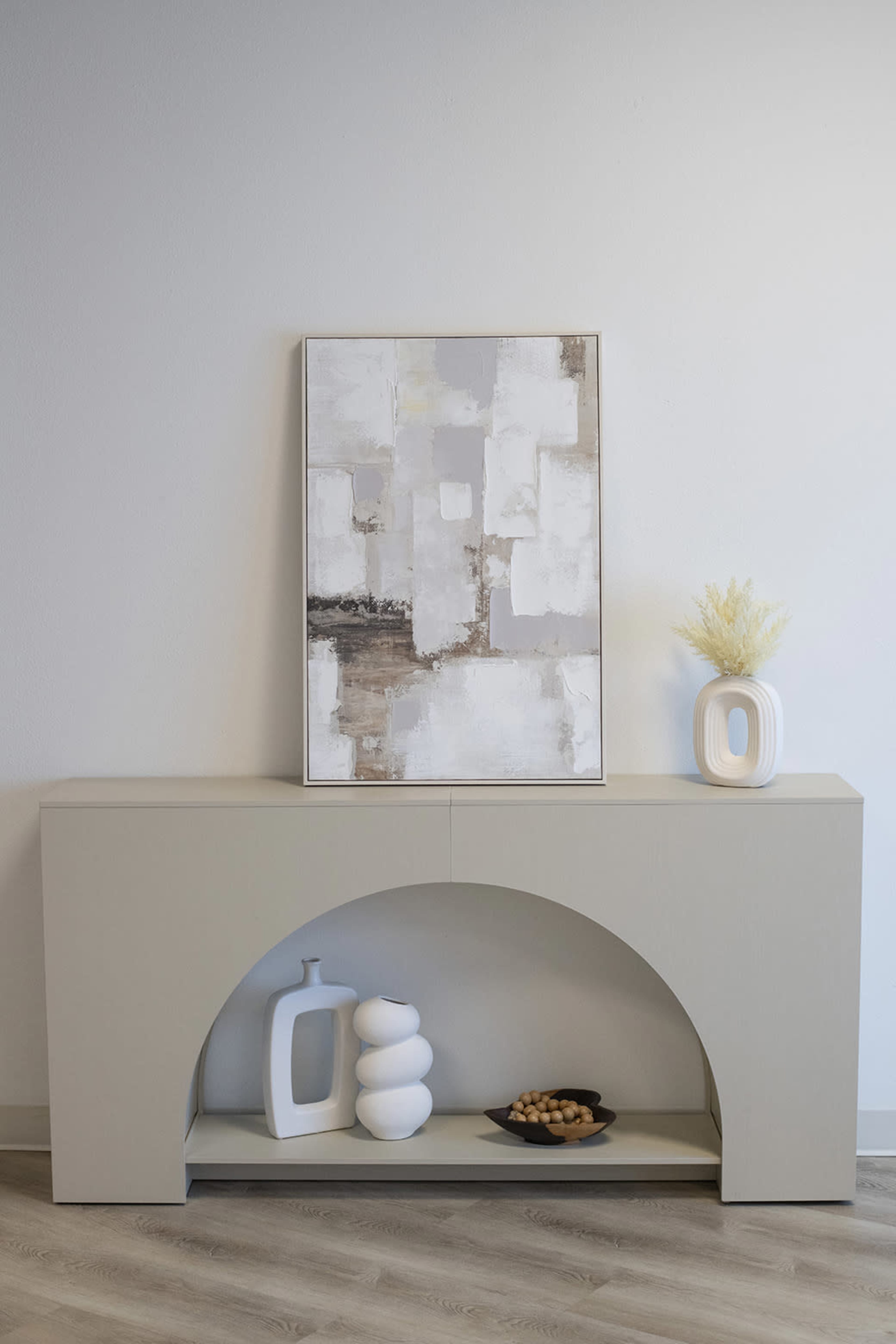 A light-colored console table with an arch design displays a framed abstract painting and decorative objects, including a vase and a bowl with small decorative items.