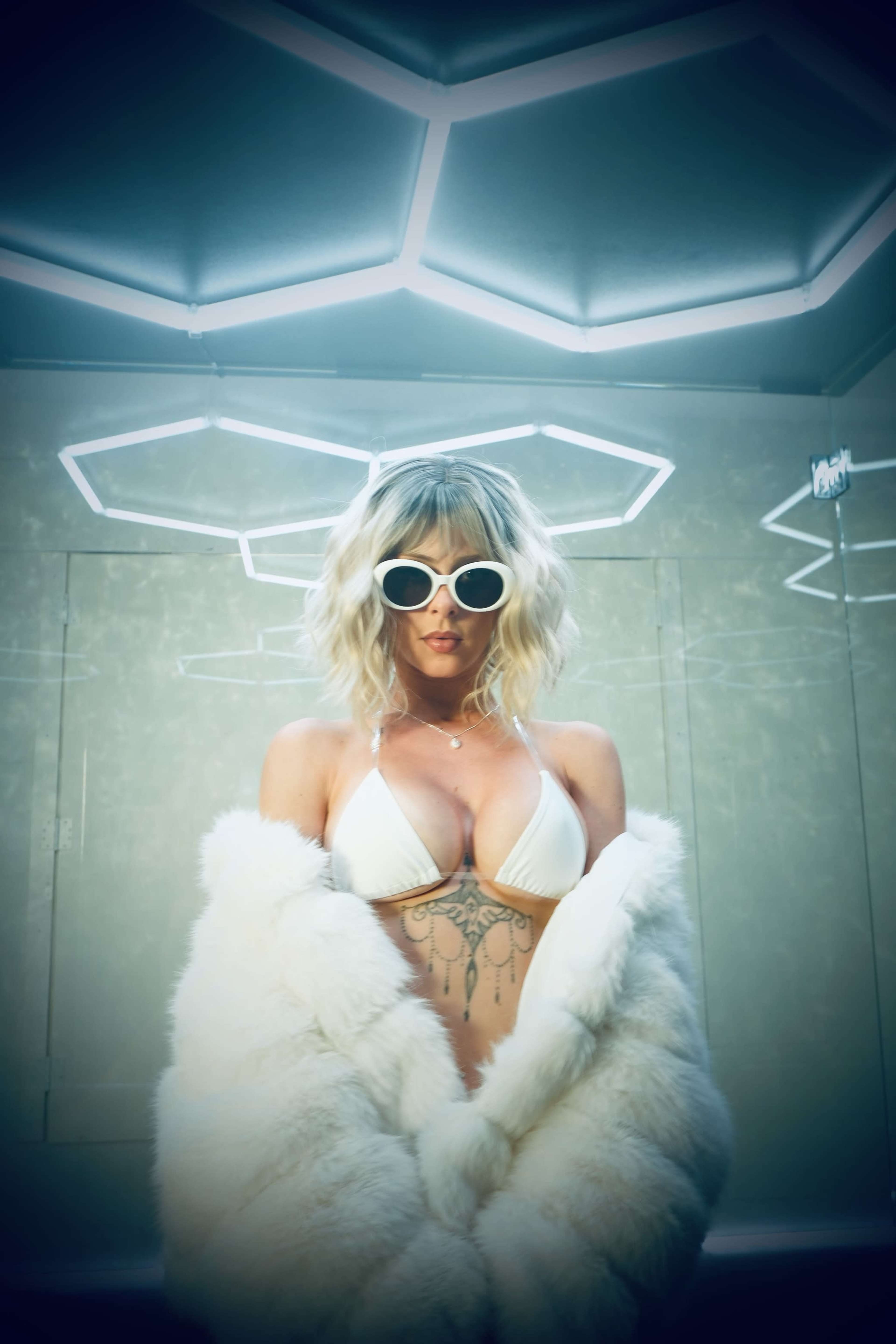 A person poses in a white bikini and fur coat under illuminated geometric lighting.