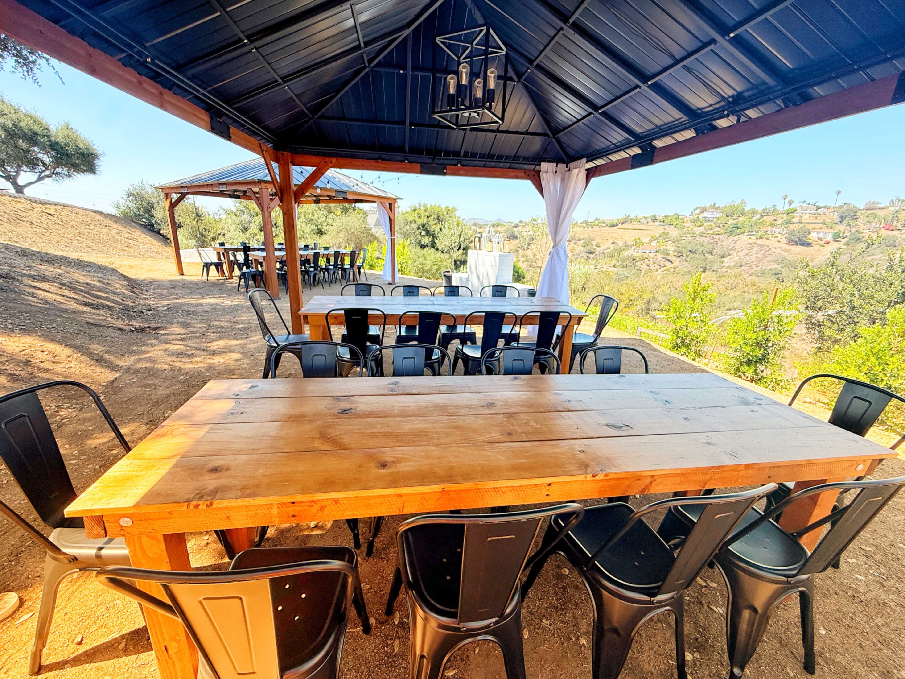 A large wooden table surrounded by black metal chairs is set under a gazebo, overlooking a hilly landscape with greenery.