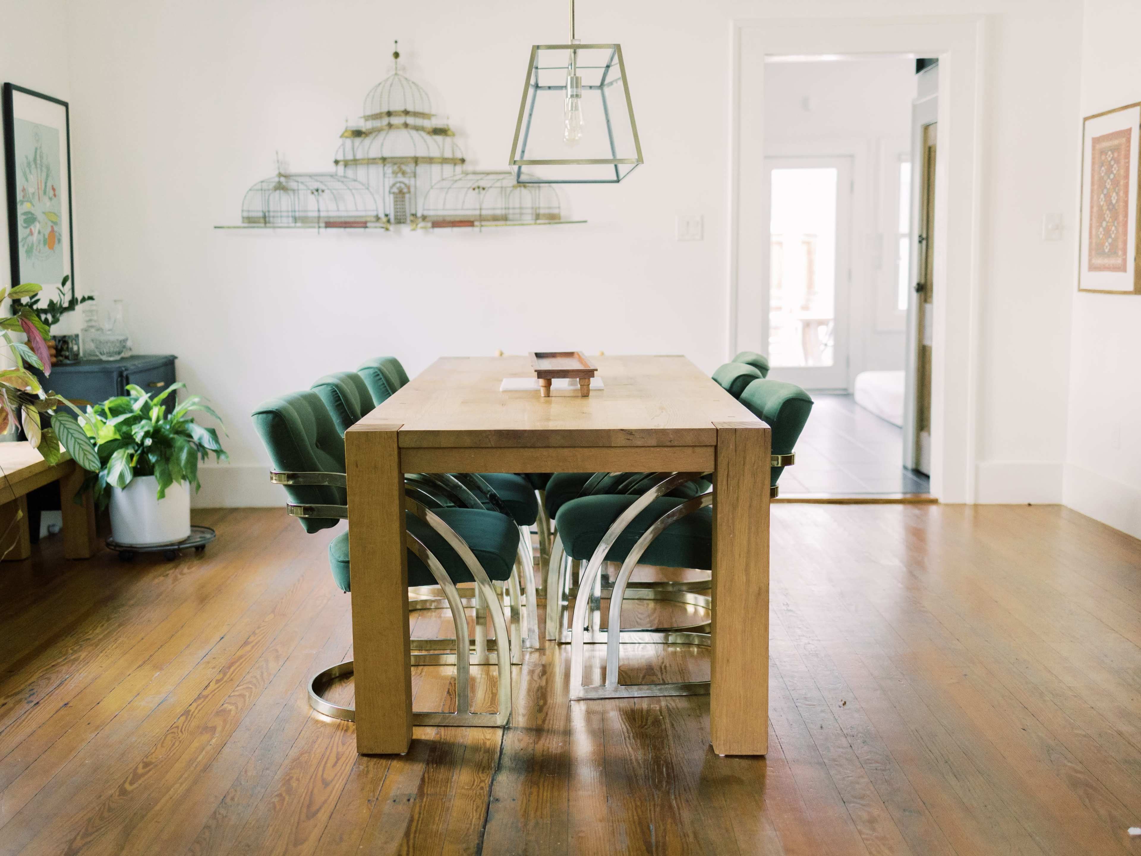 A wooden dining table with metallic legs is surrounded by green upholstered chairs in a well-lit room featuring a doorway and decorative items.