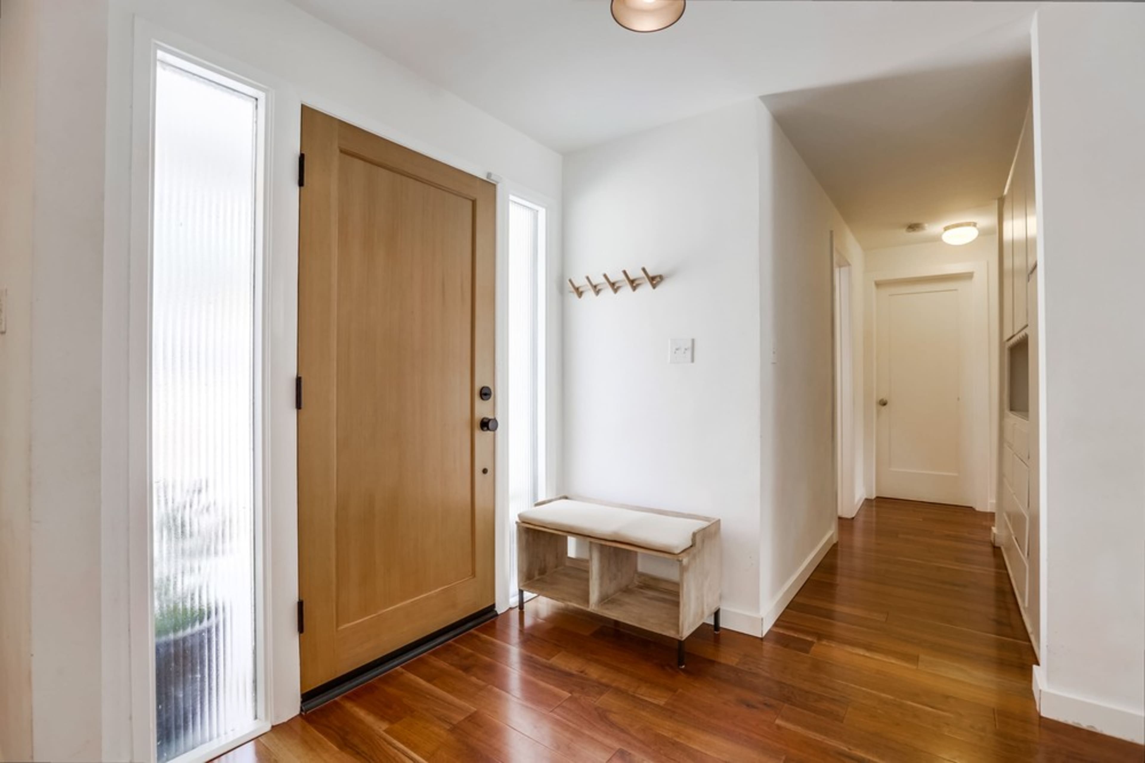A minimalist entryway featuring a wooden front door, a light-colored bench, and hardwood flooring, with windows on both sides of the door.