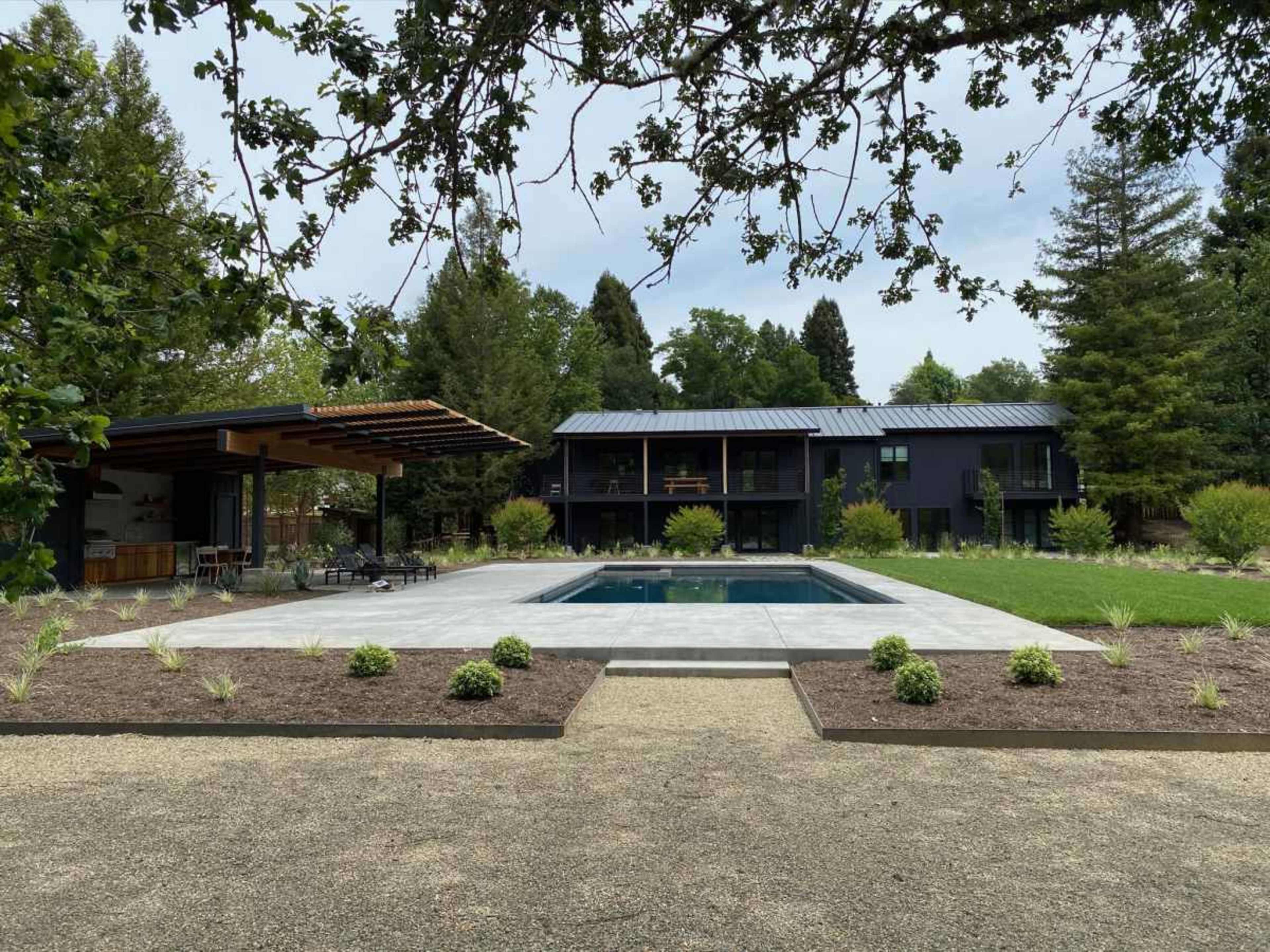 The image shows a modern, two-story black house with a swimming pool in the foreground and a covered outdoor seating area nearby, surrounded by landscaped greenery.