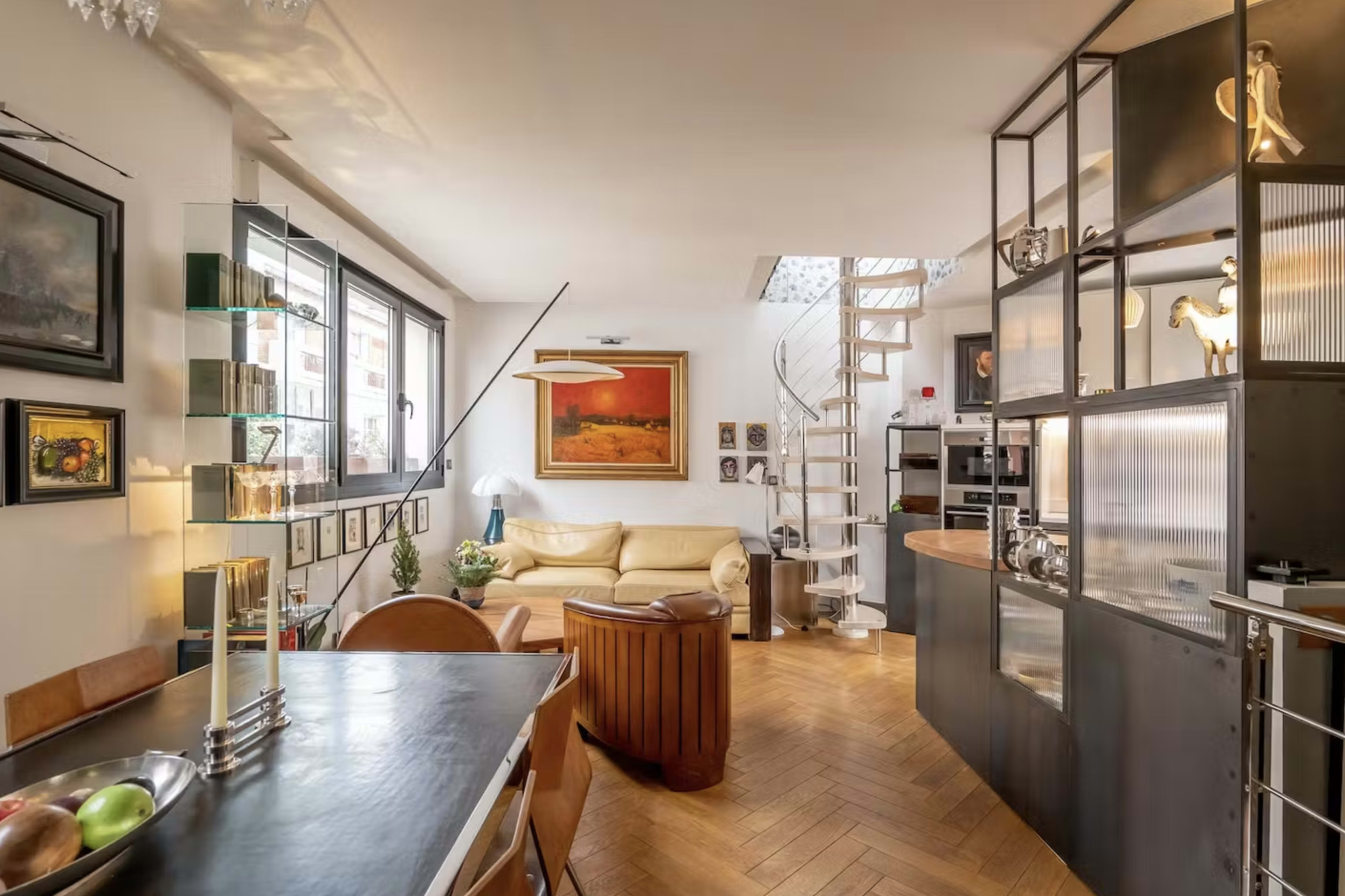 The image shows a modern living space with an open layout that includes a dining area, a lounge with a yellow sofa, and a kitchen with a spiral staircase in the background.