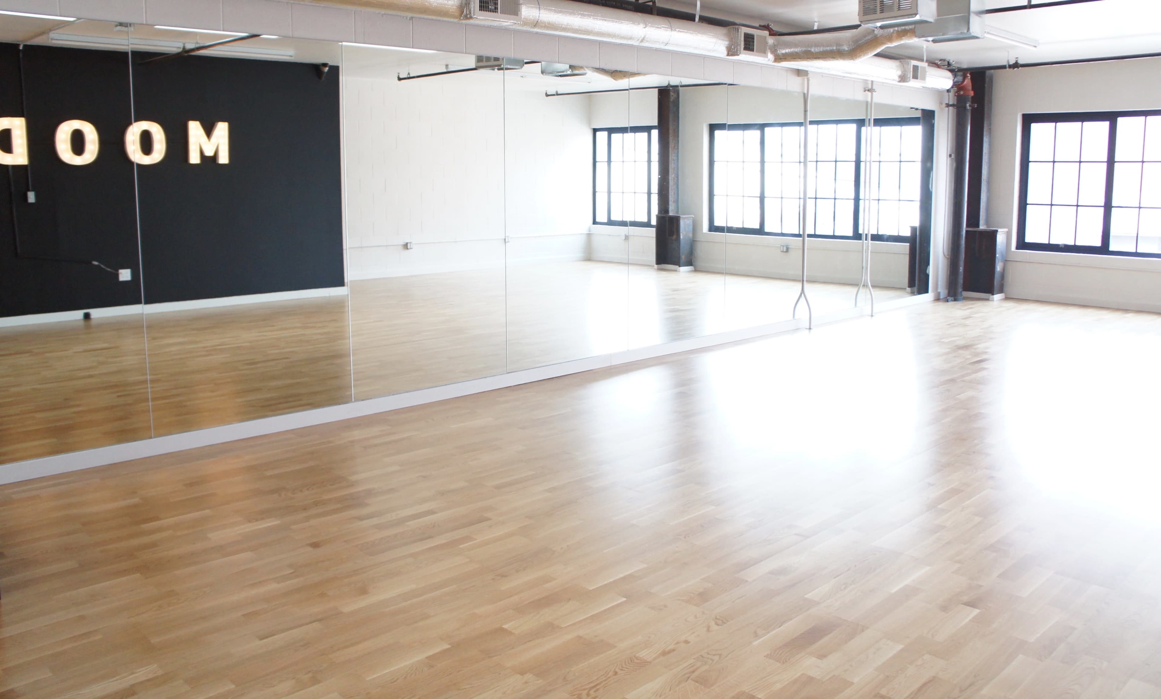 A bright, empty dance studio featuring wooden floors, large mirrors, and windows.