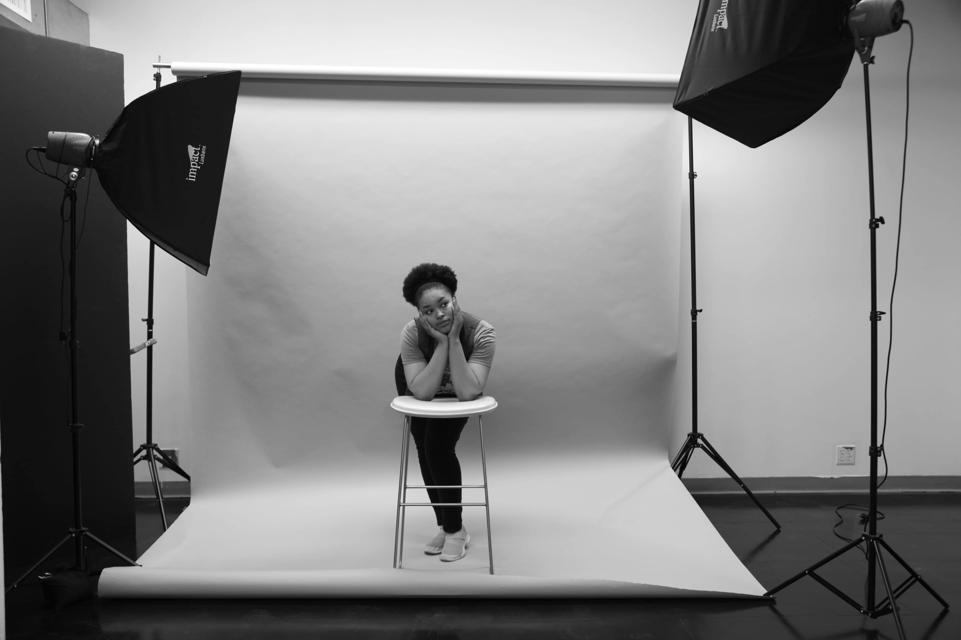 A person sits on a stool in a photography studio with soft lighting and a neutral backdrop.