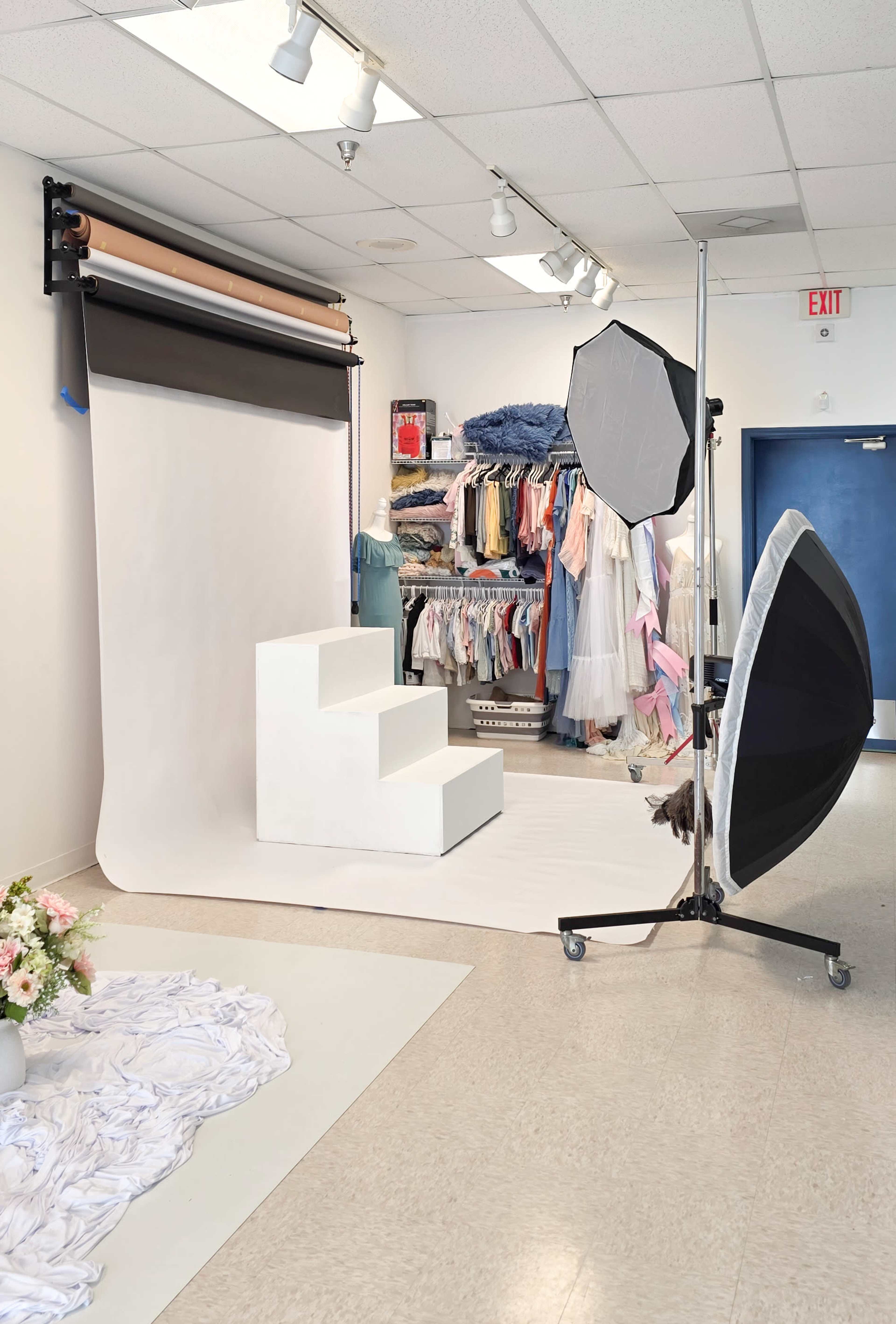 The image shows a photography studio with a white backdrop, a three-step platform, lighting equipment, and a clothing rack filled with various garments.