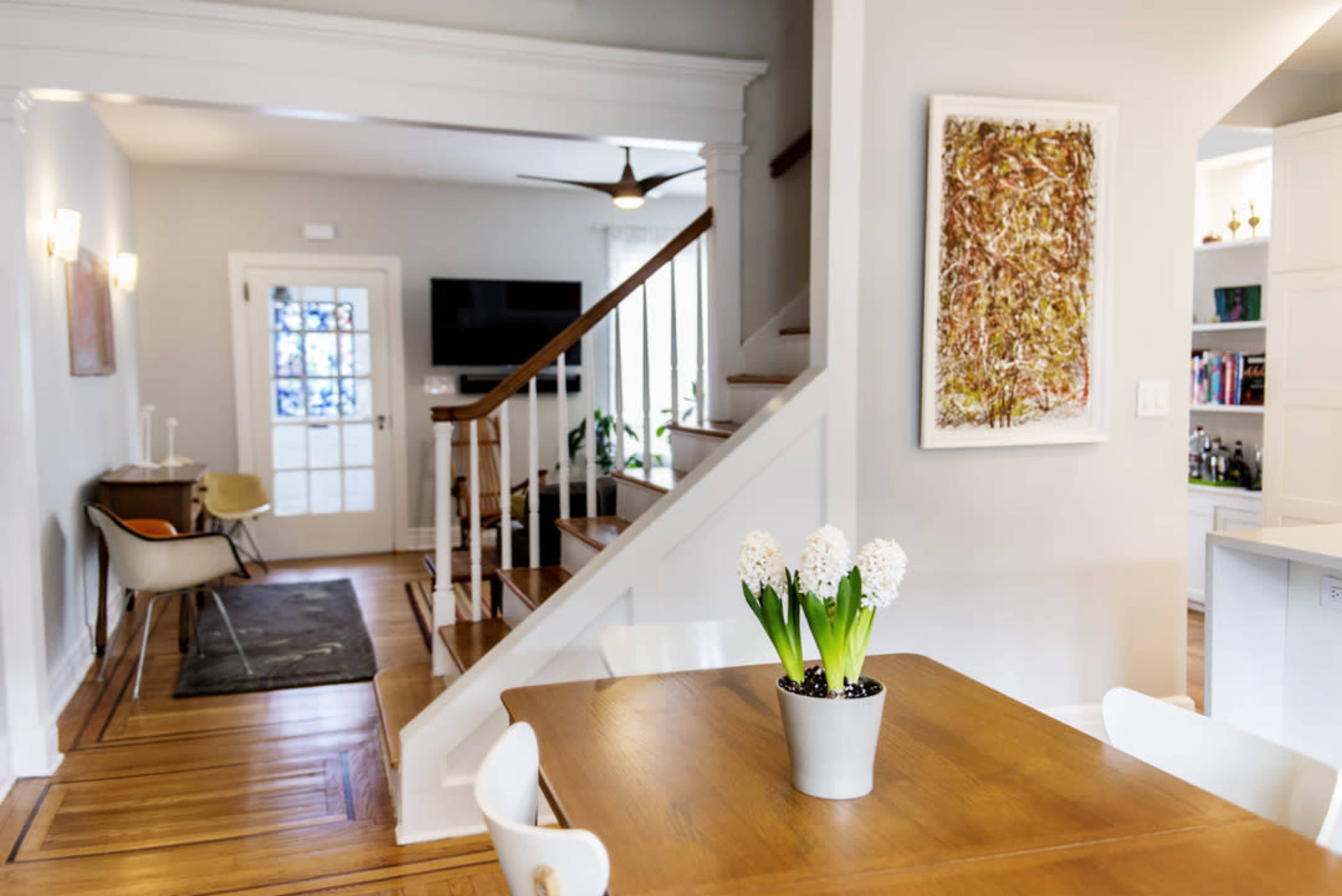A well-lit living space features a wooden dining table with a potted plant, adjacent to a staircase and a cozy seating area.
