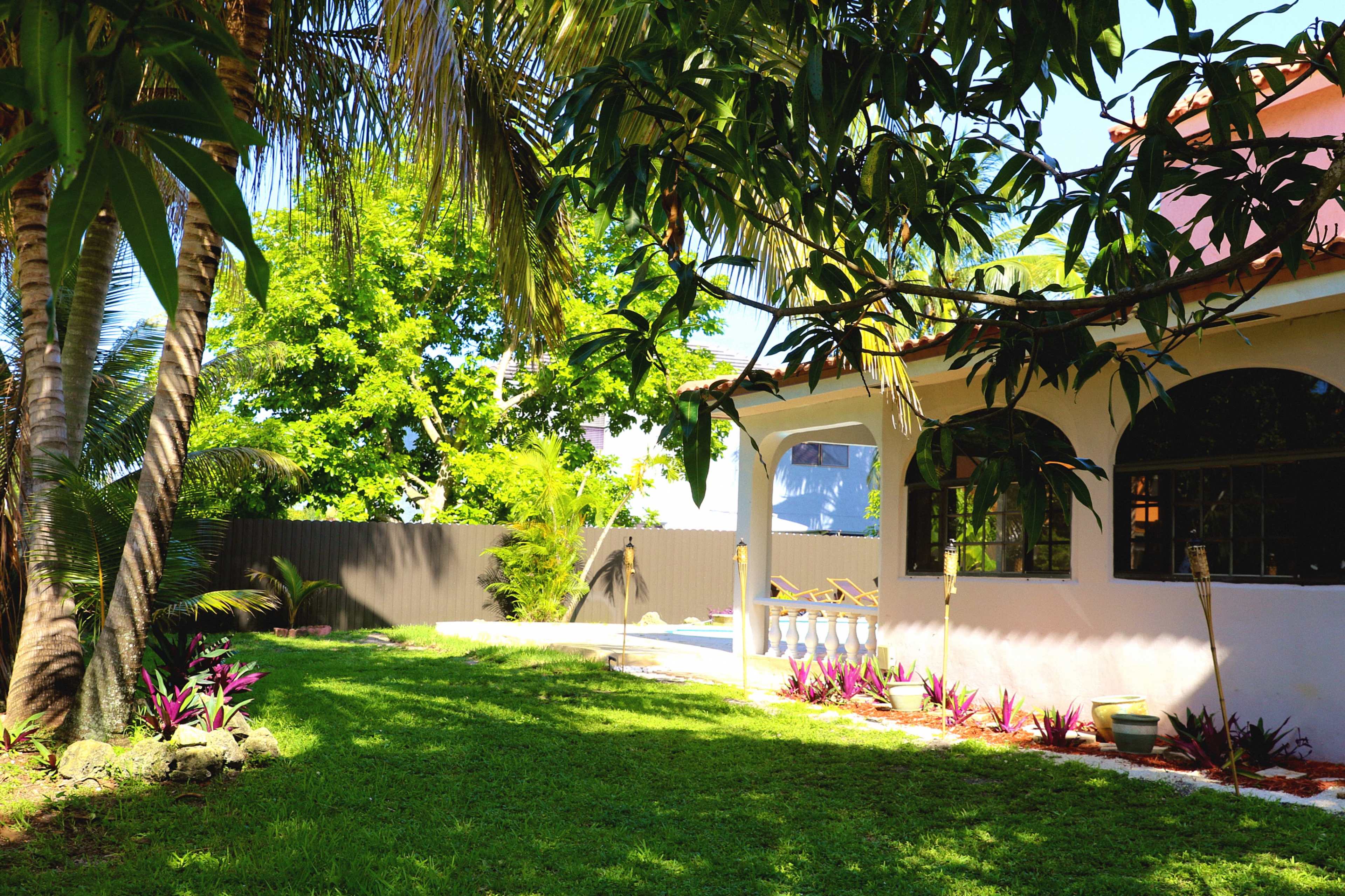 A well-maintained garden features green grass, tropical plants, and a house with arched windows and a patio.
