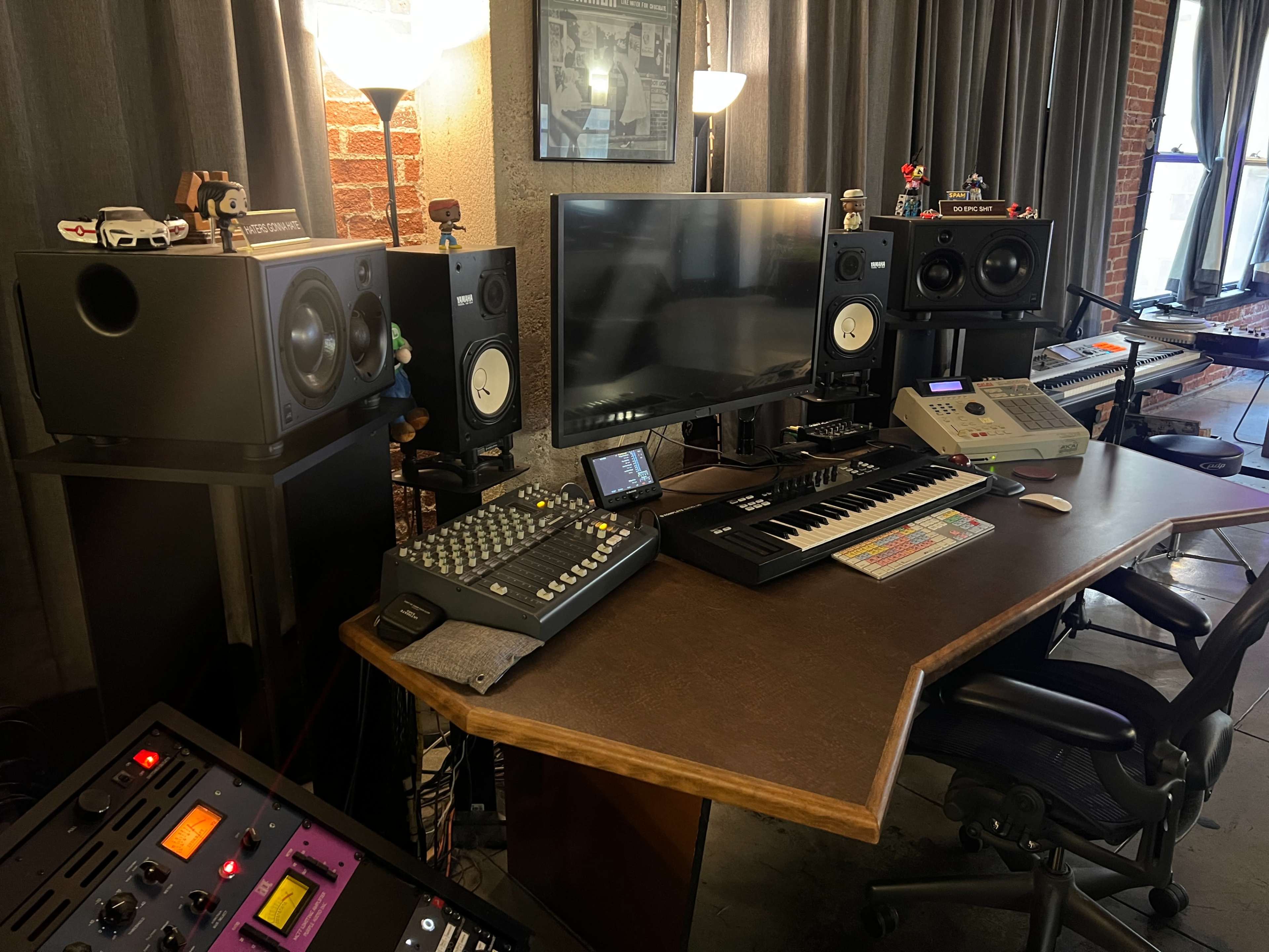 The image shows a modern music studio setup featuring a computer monitor, keyboards, audio speakers, a mixing console, and various music production equipment arranged around a wooden desk.