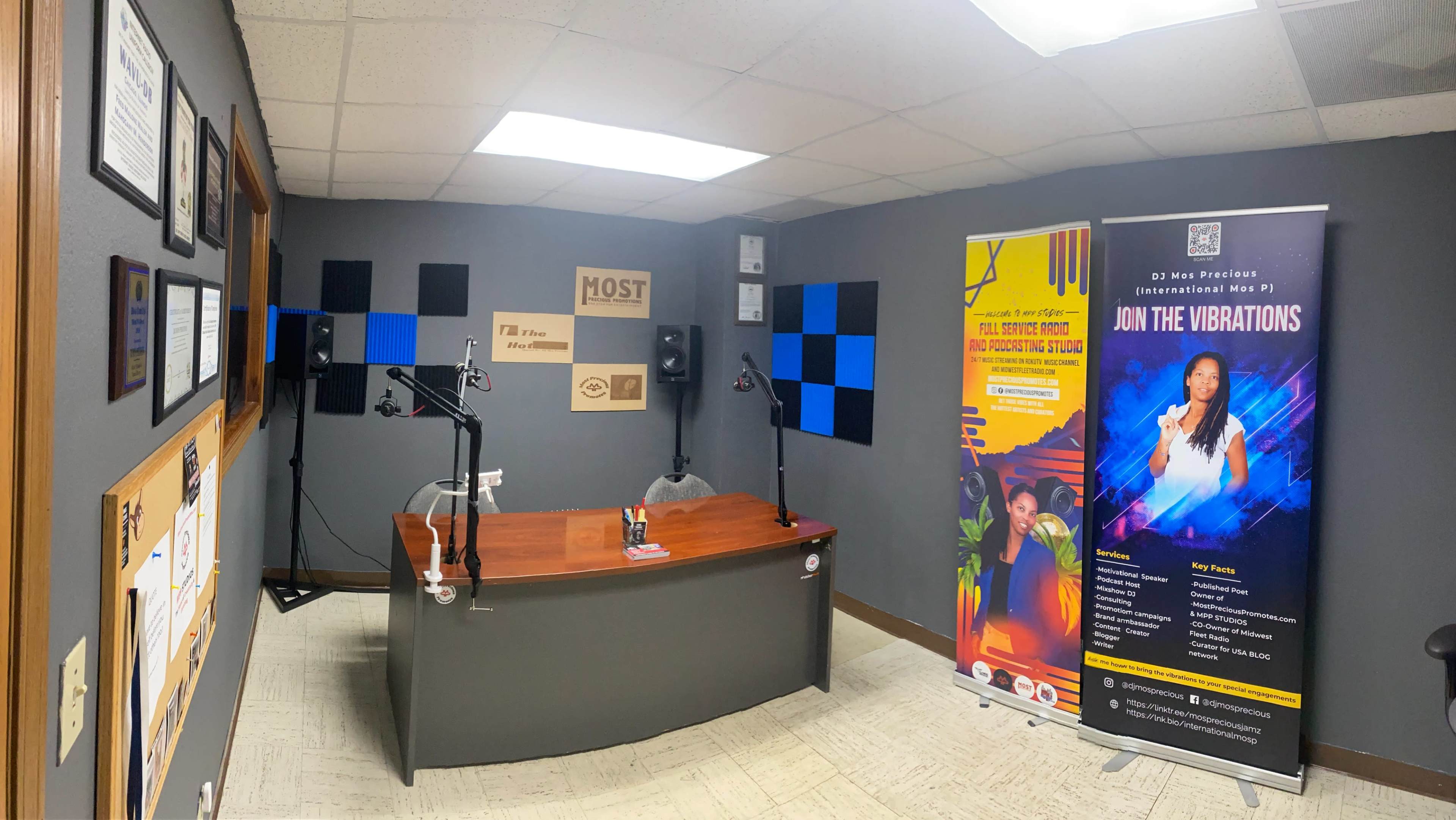 A radio studio featuring a wooden desk with microphones, soundproofing panels on the walls, and promotional banners on either side.