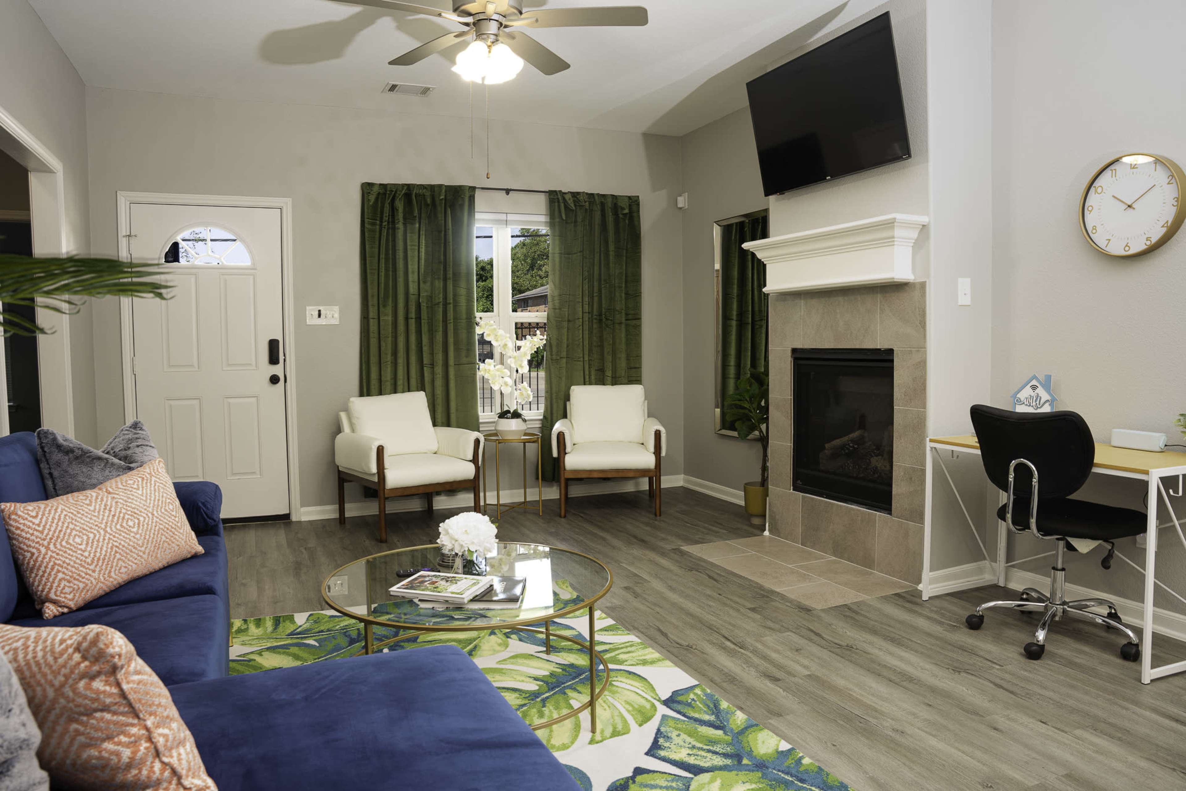 The living room features a blue sofa, two white chairs, a coffee table, a fireplace, and a desk, with green curtains and large windows allowing natural light.