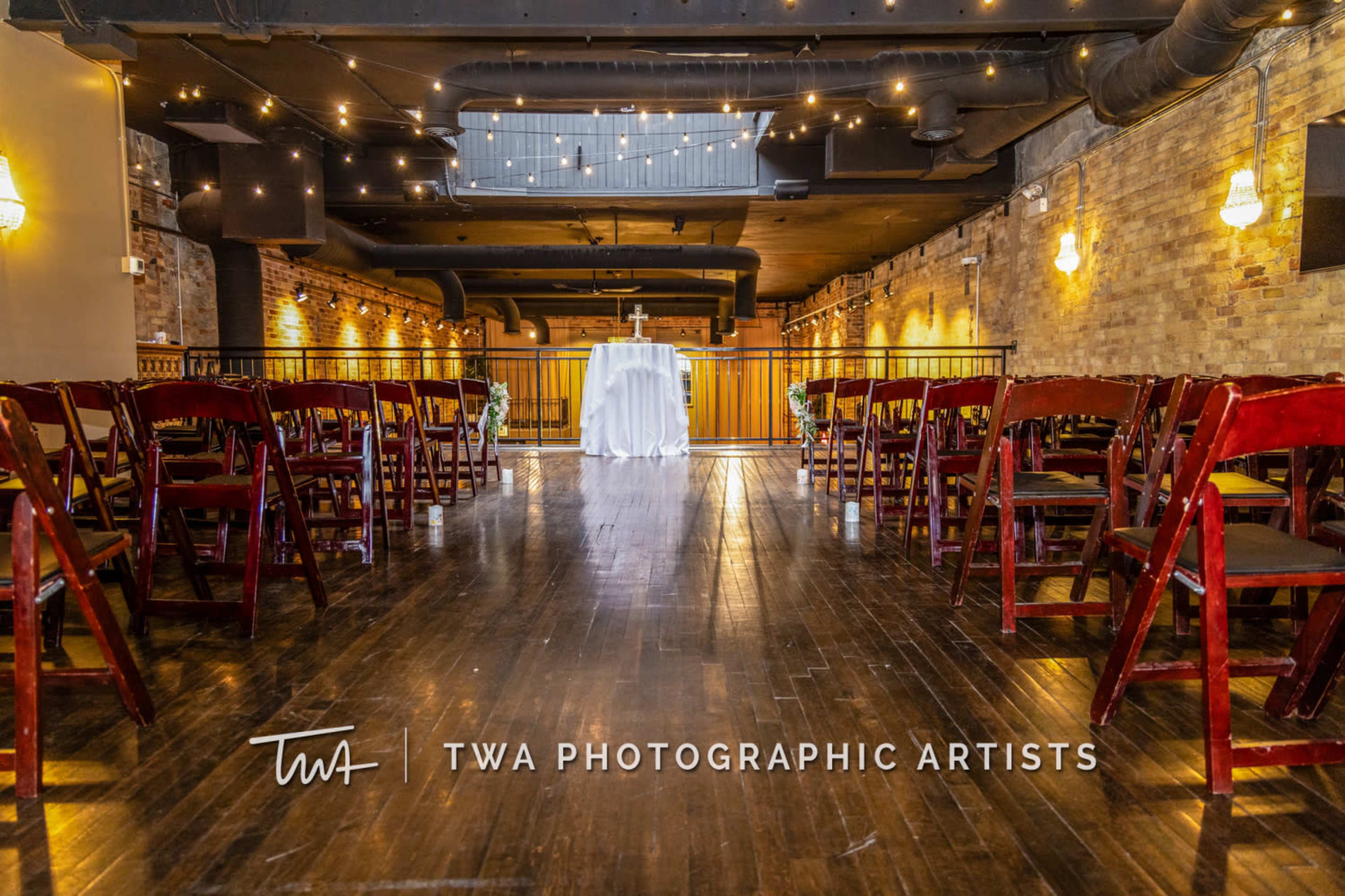 The image shows an empty event space with rows of wooden chairs facing a table at the front, under warm lighting and exposed brick walls.