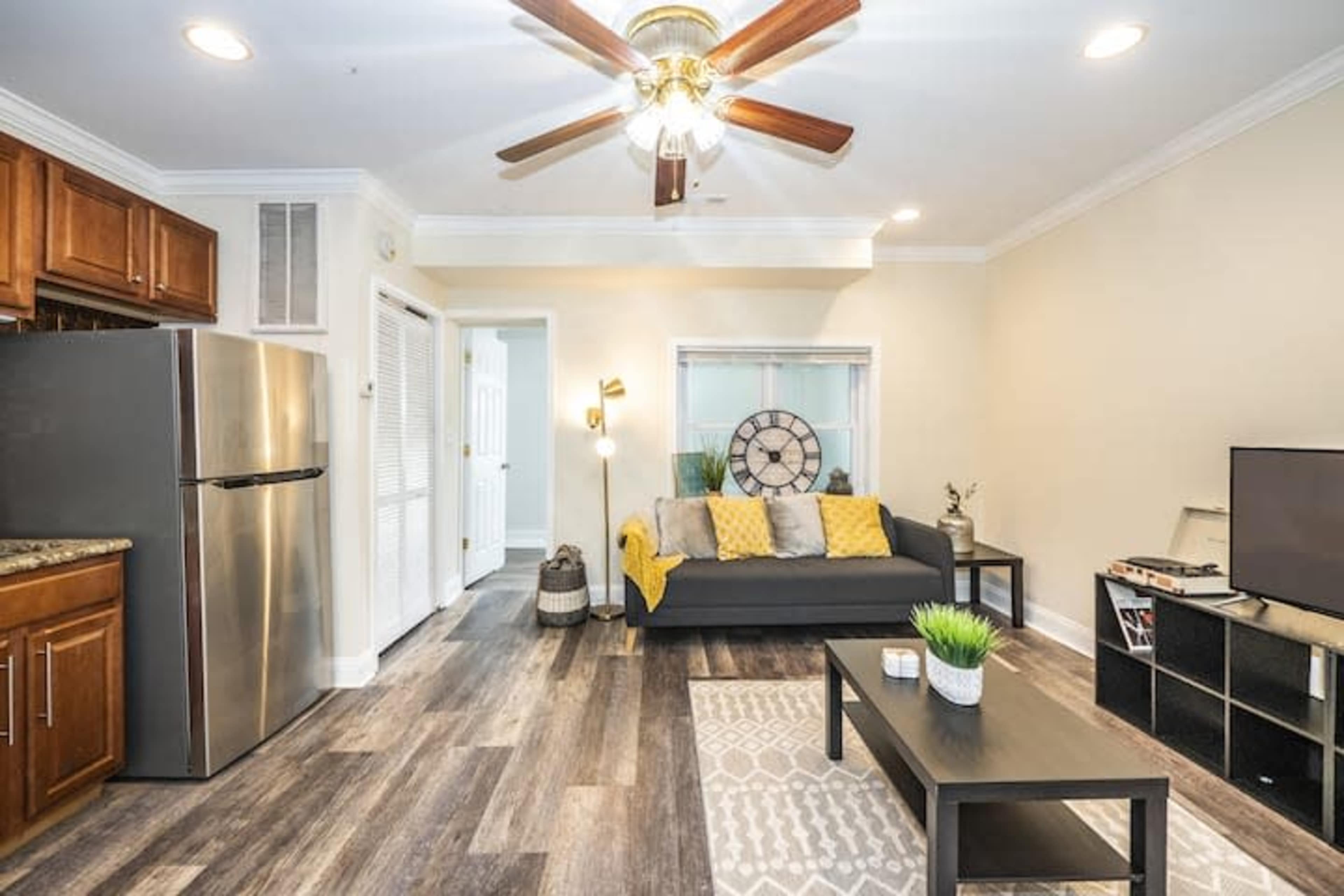 A living space with a couch, a coffee table, a television, and a kitchen area featuring a stainless steel refrigerator and wooden cabinets.