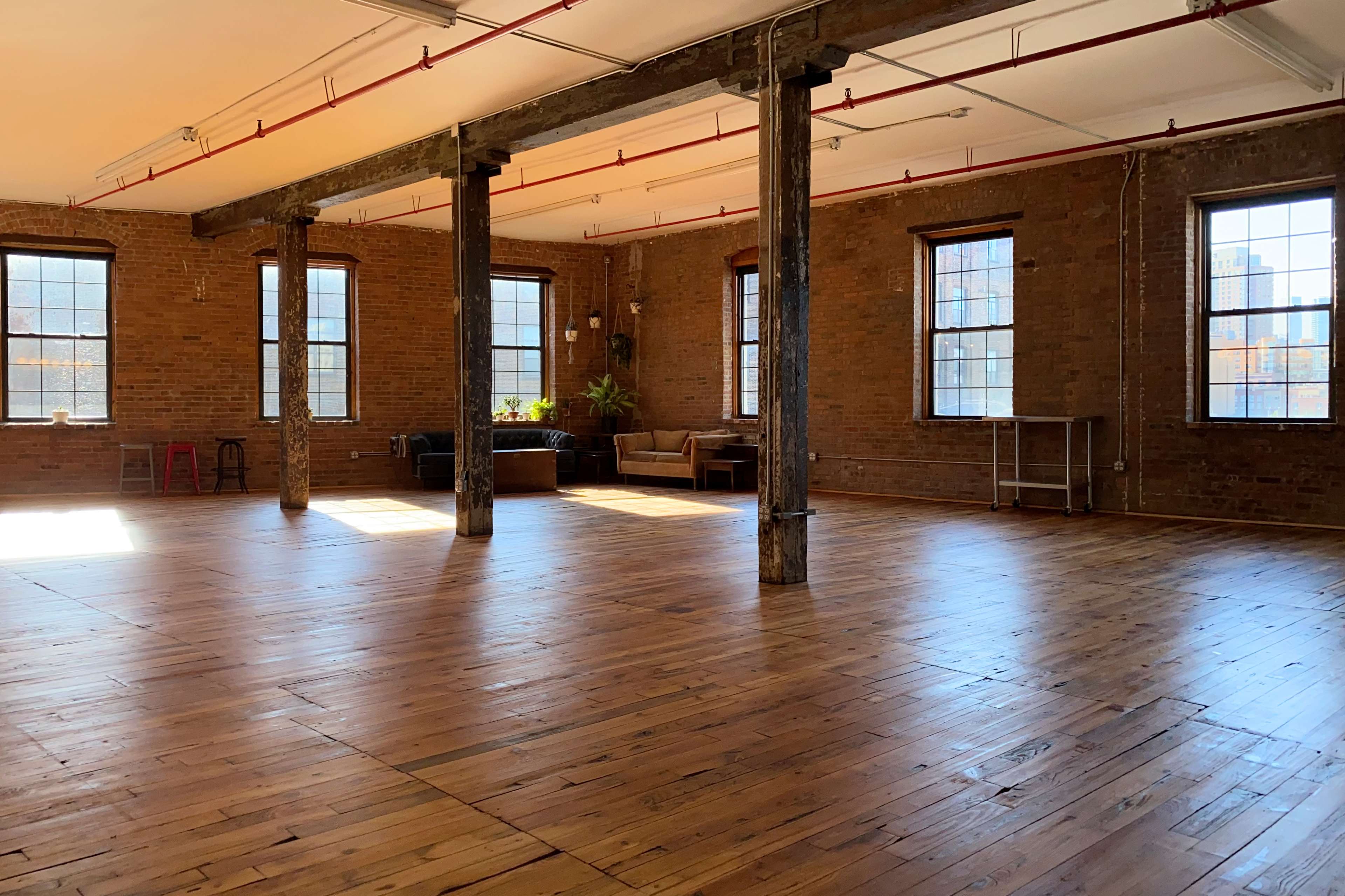 The image shows an empty industrial-style room with wooden floors, brick walls, and large windows offering natural light.