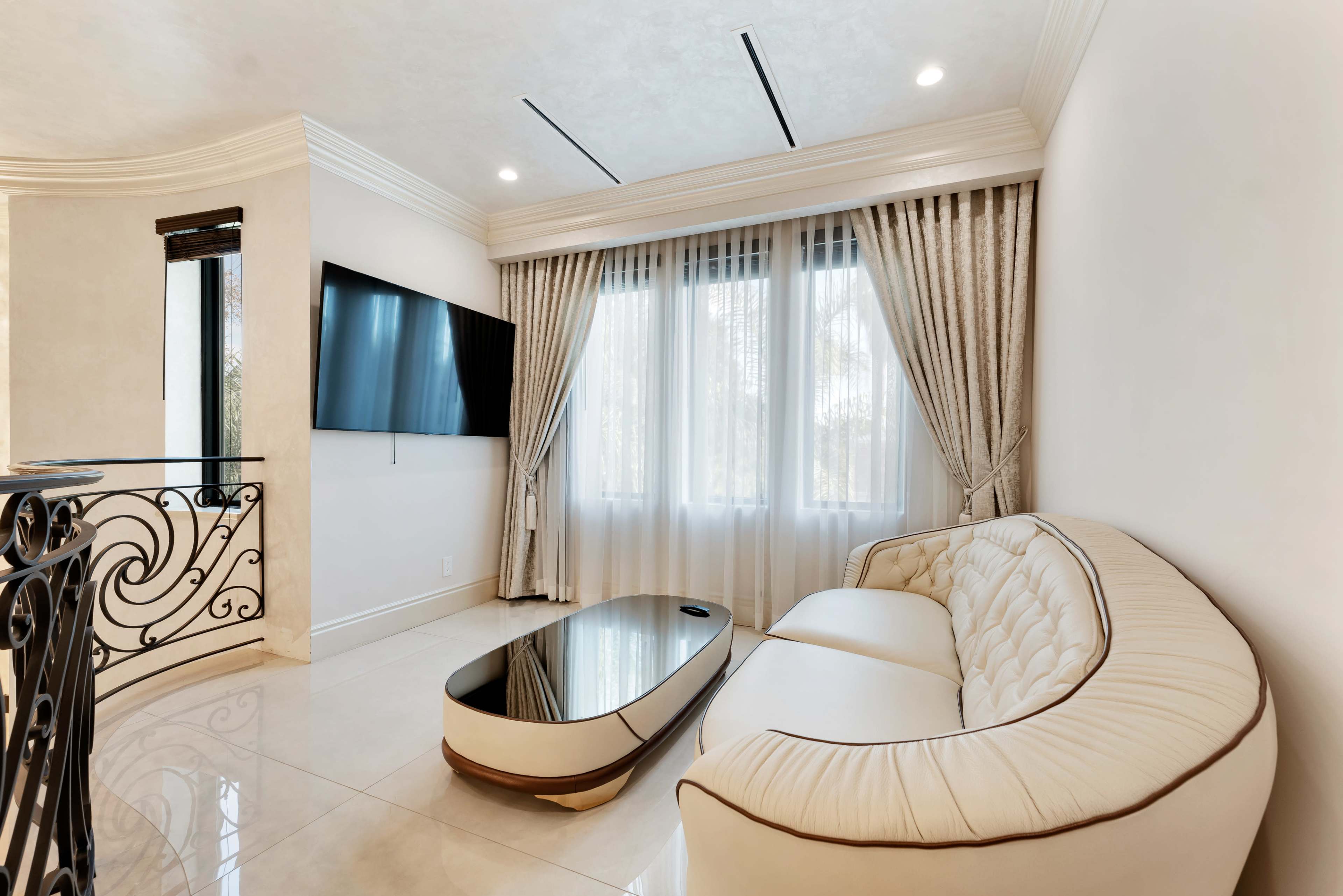 The image shows a modern living room with a white sofa, a glass coffee table, and large windows covered by sheer curtains.