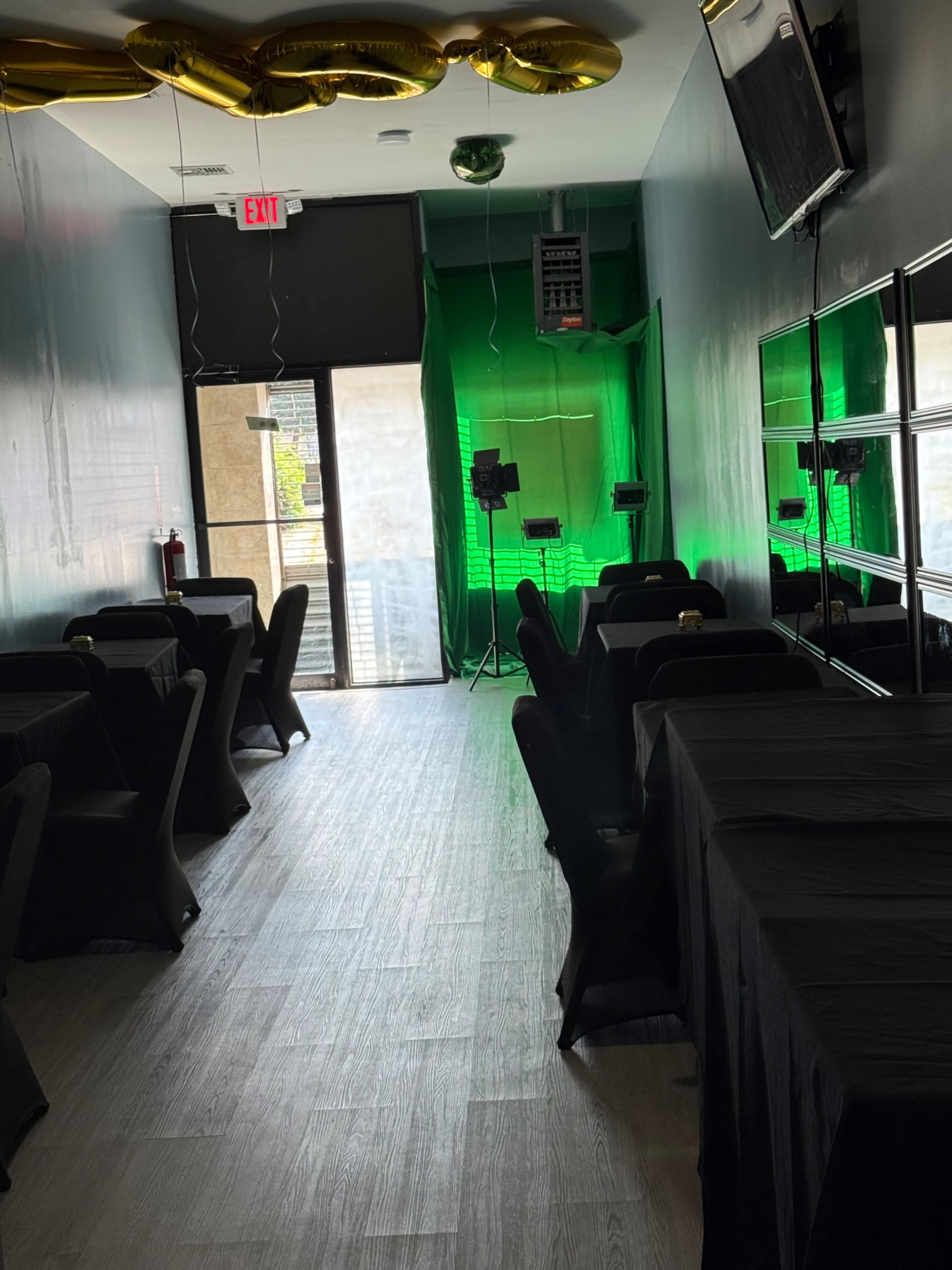 A narrow, dimly lit room features black tablecloths on several tables, with green drapery and lighting at the far end.